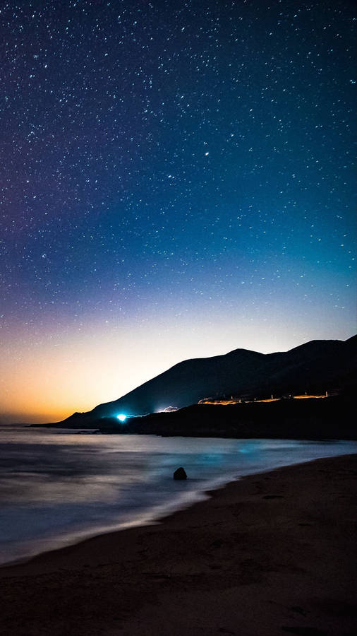 繁星點點的夜晚，海灘上有山丘和沿岸的遠方燈光。 — 4K Android 壁紙