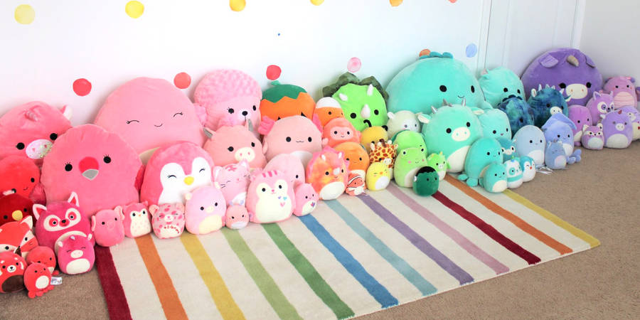 一張彩虹地毯，上面有一排色彩繽紛的毛絨玩具，背景為圓點牆。 — Squishmallows 壁紙