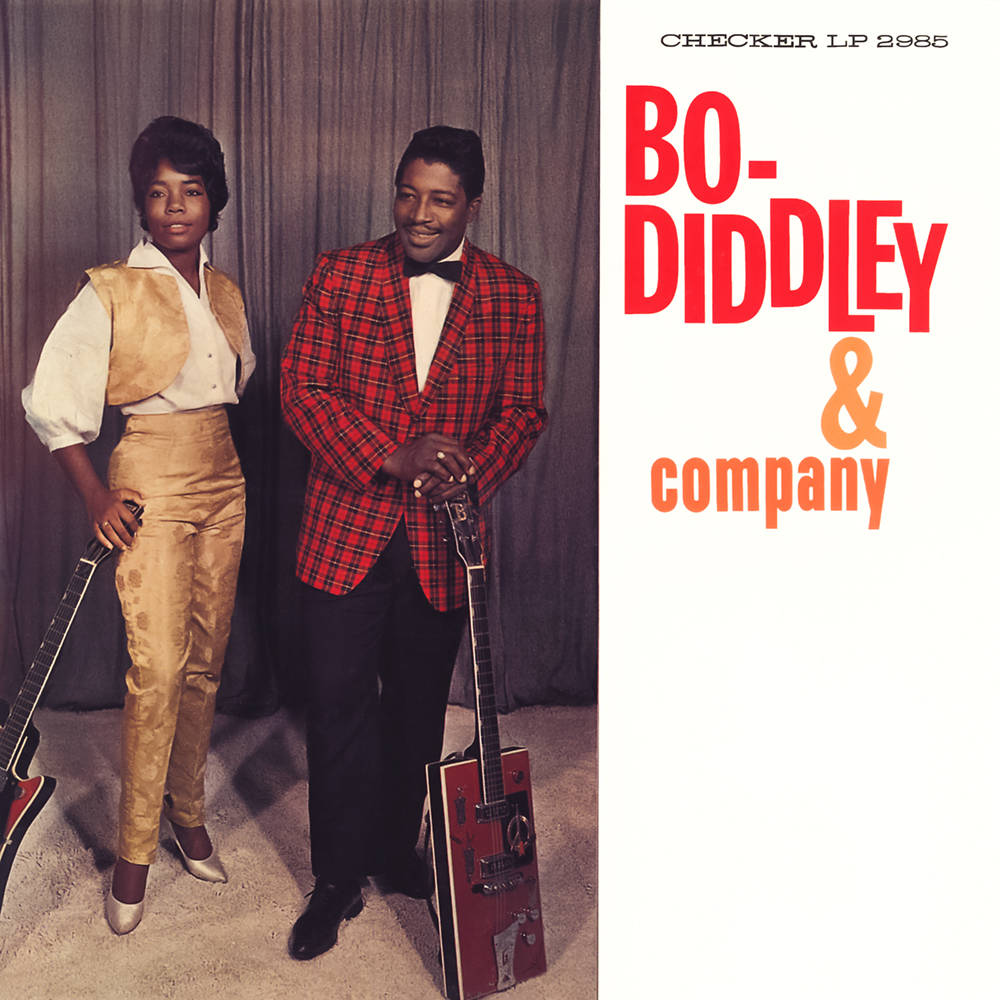 吉他動圖 桌布 壁紙 — 1960年代復古照片，Bo Diddley 與一名身穿大膽格紋和金色服裝的女子，在窗簾背景下手持吉他。