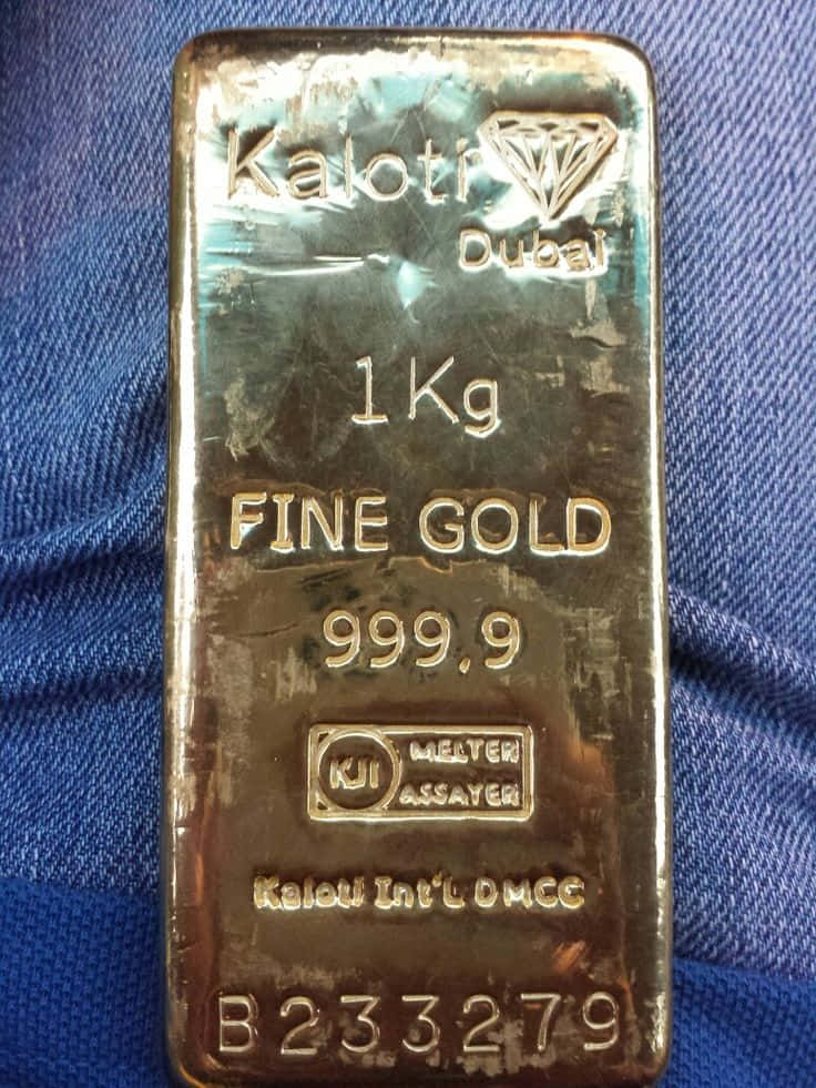 Gold Bars 桌布 壁紙 — 1公斤Kaloti Dubai純金金條的特寫，放置在藍色牛仔布上，顯示雕刻標記和金屬光澤。