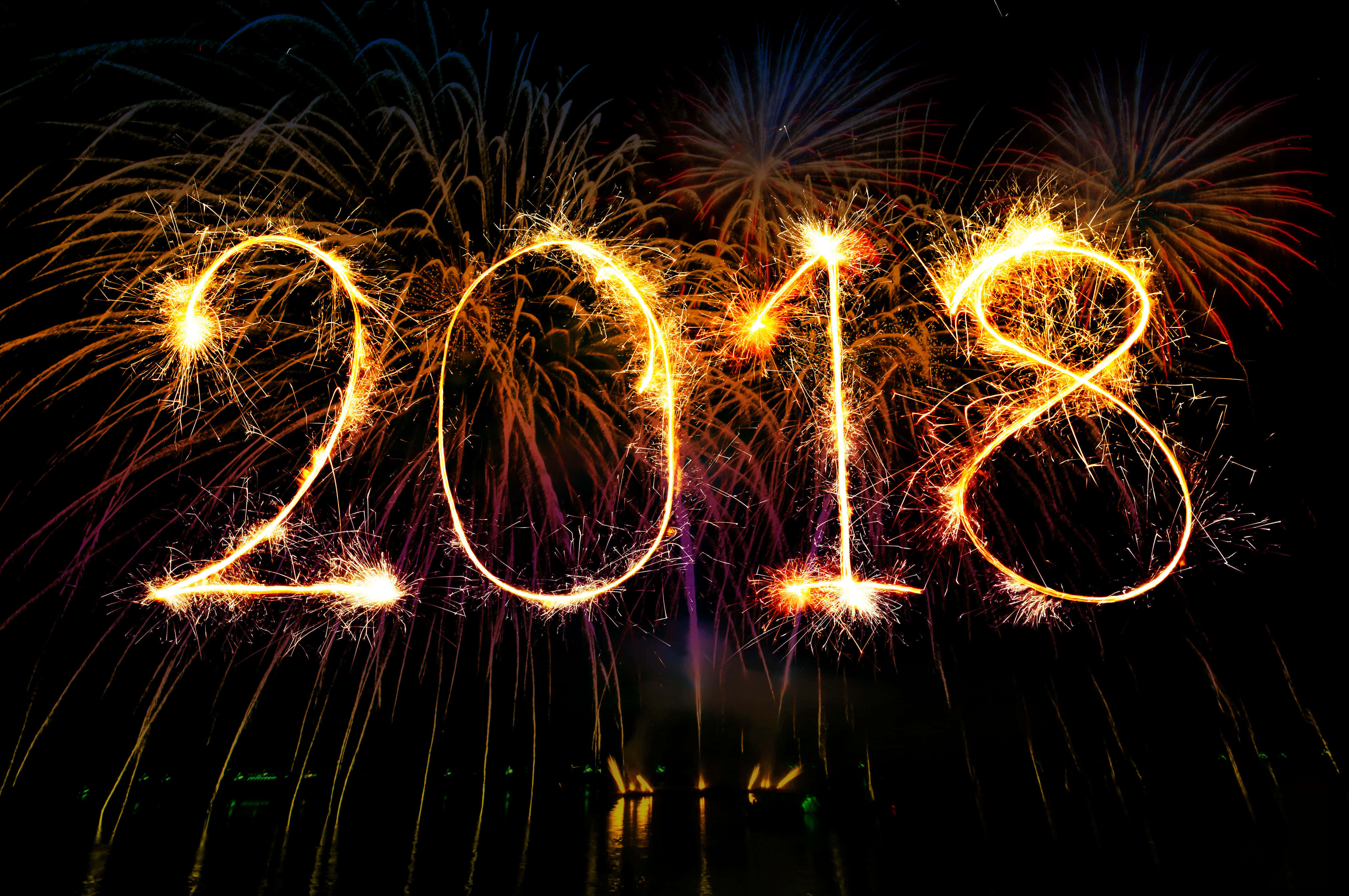 2018 桌布 壁紙 — 2018年以發光的金色煙火棒拼寫，背景是黑暗的夜空，後面有色彩繽紛的煙火爆炸。