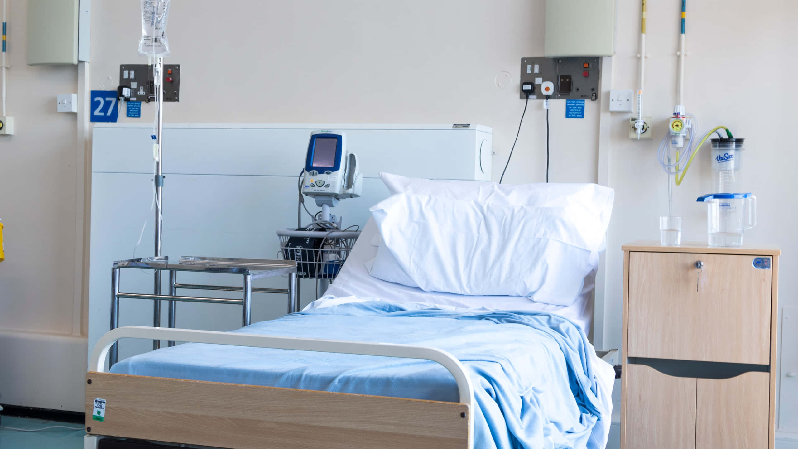 Hospital 桌布 壁紙 — 27號房內一張乾淨的醫院病床，配有醫療設備和藍色床單，在明亮的螢光燈下。