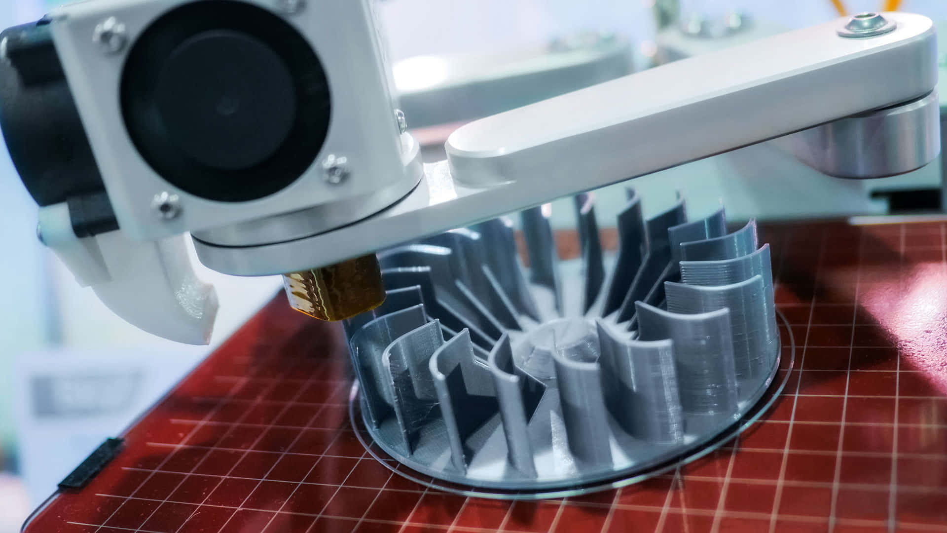 3d Printer 桌布 壁紙 — 3D列印機特寫，正在擠出灰色線材，在紅色網格床上構建一個風扇形狀的物體。