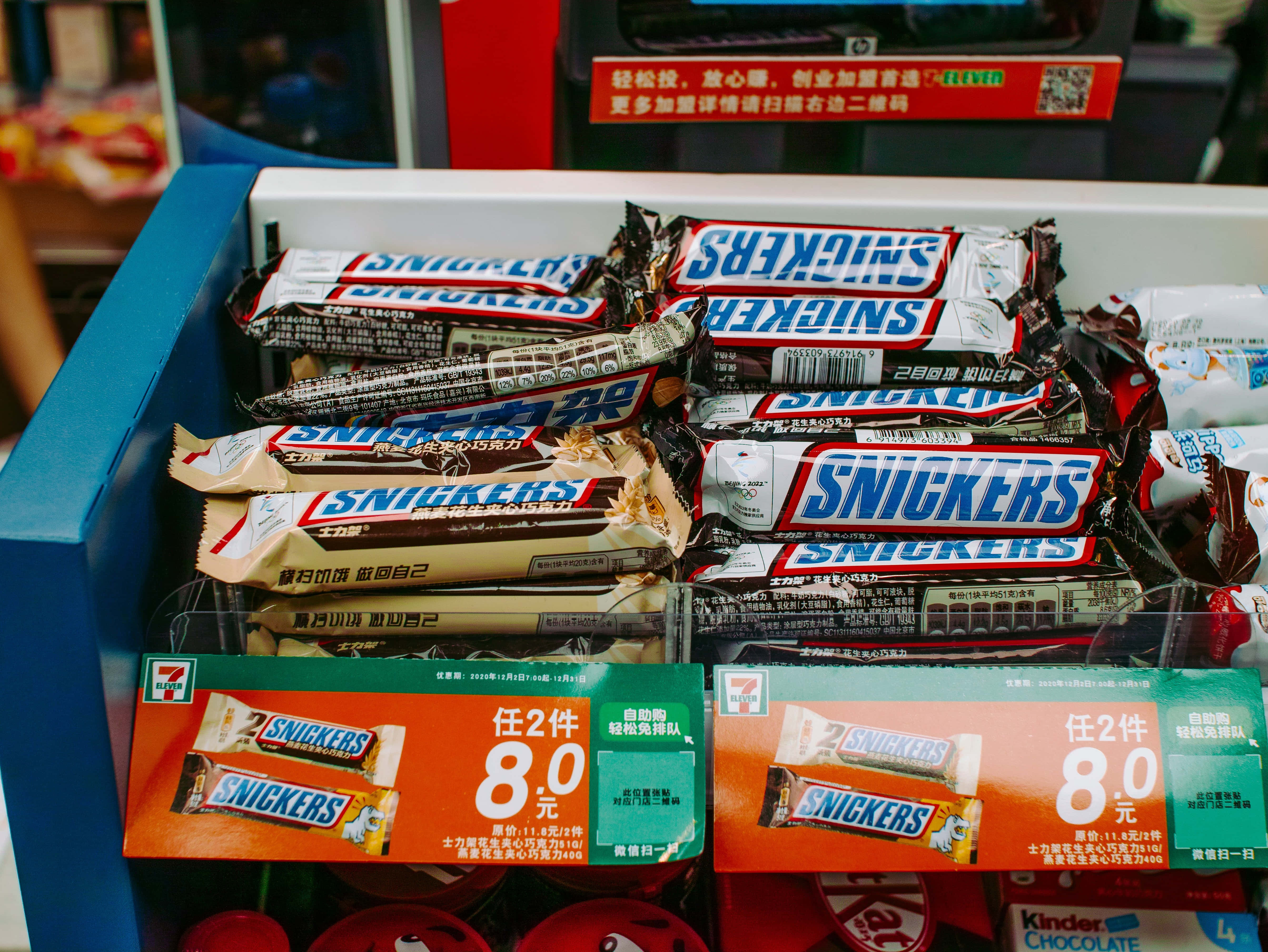 高清 桌布 壁紙 — 7-Eleven 店內展示架上堆疊的 Snickers 巧克力棒特寫，附有鮮橙色和綠色的價格標籤。