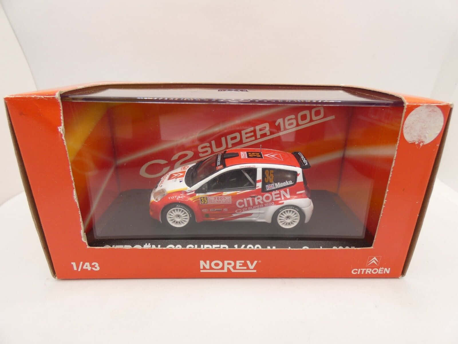 Citroen C2 Super 1600 桌布 壁紙 — 一個1:43比例的雪鐵龍C2 Super 1600拉力賽車模型，採用紅、白、黑配色，展示在其橙色品牌盒中。