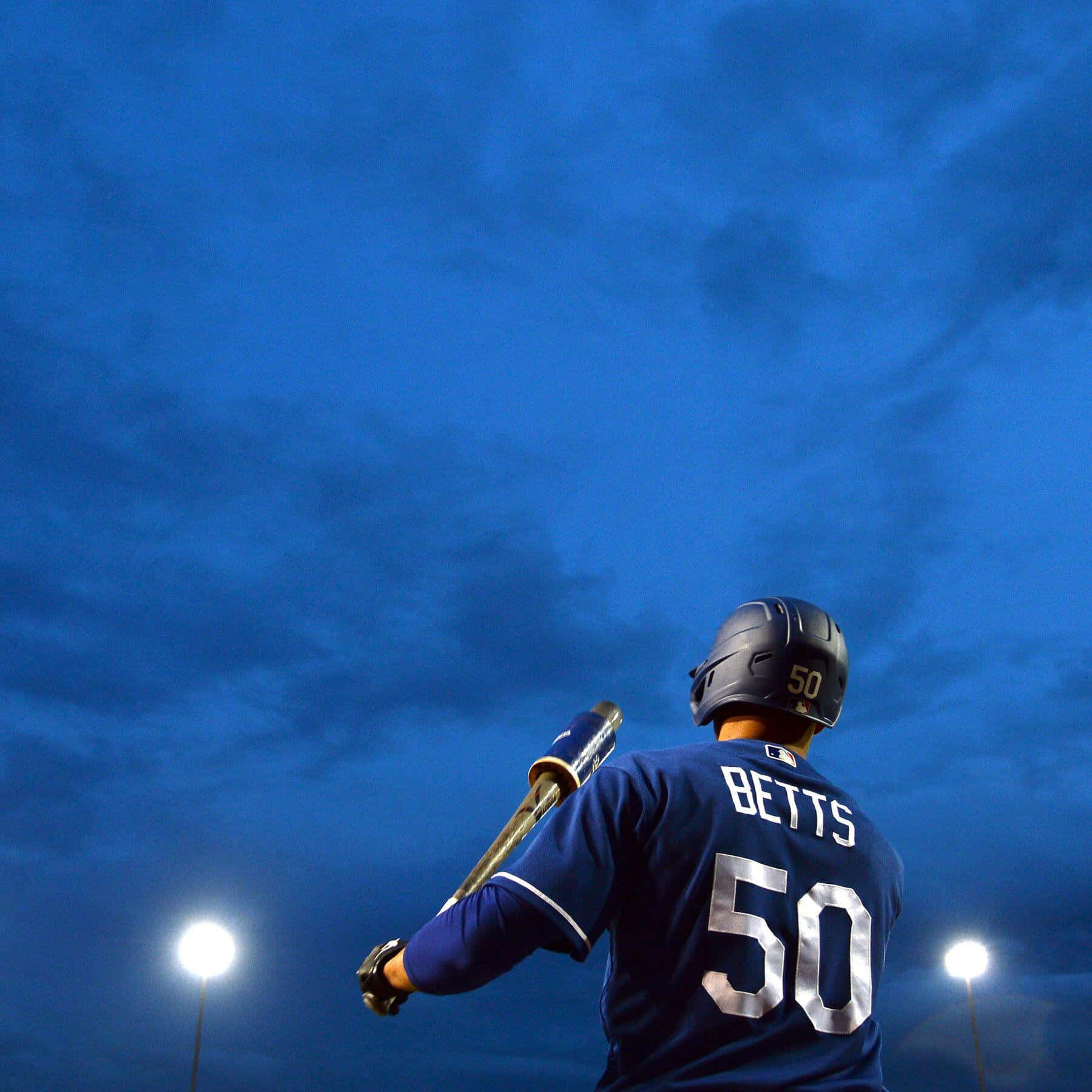  壁紙 — 一名身穿印有BETTS 50的藍色球衣的棒球運動員，在深藍色暮色天空的背景下，站在體育場燈光下。