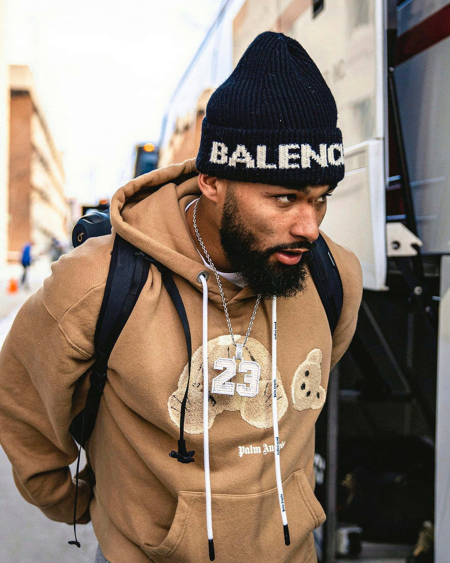  壁紙 — 一位留鬍鬚的男子戴著黑色 Balenciaga 毛帽和米色連帽衫，背著背包走在城市街道上。
