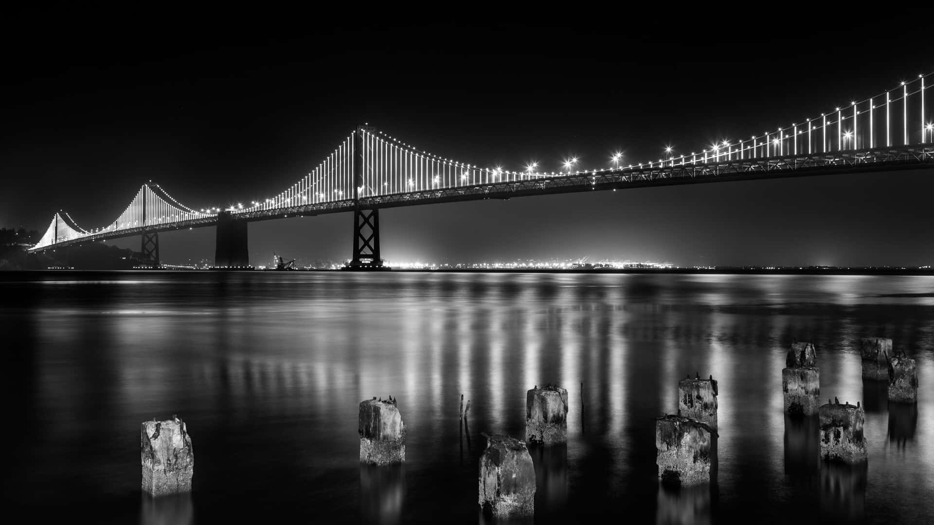  壁紙 — 一張黑白夜景照片，拍攝一座亮燈的吊橋倒映在平靜的水面上，前景有風化的樁柱。