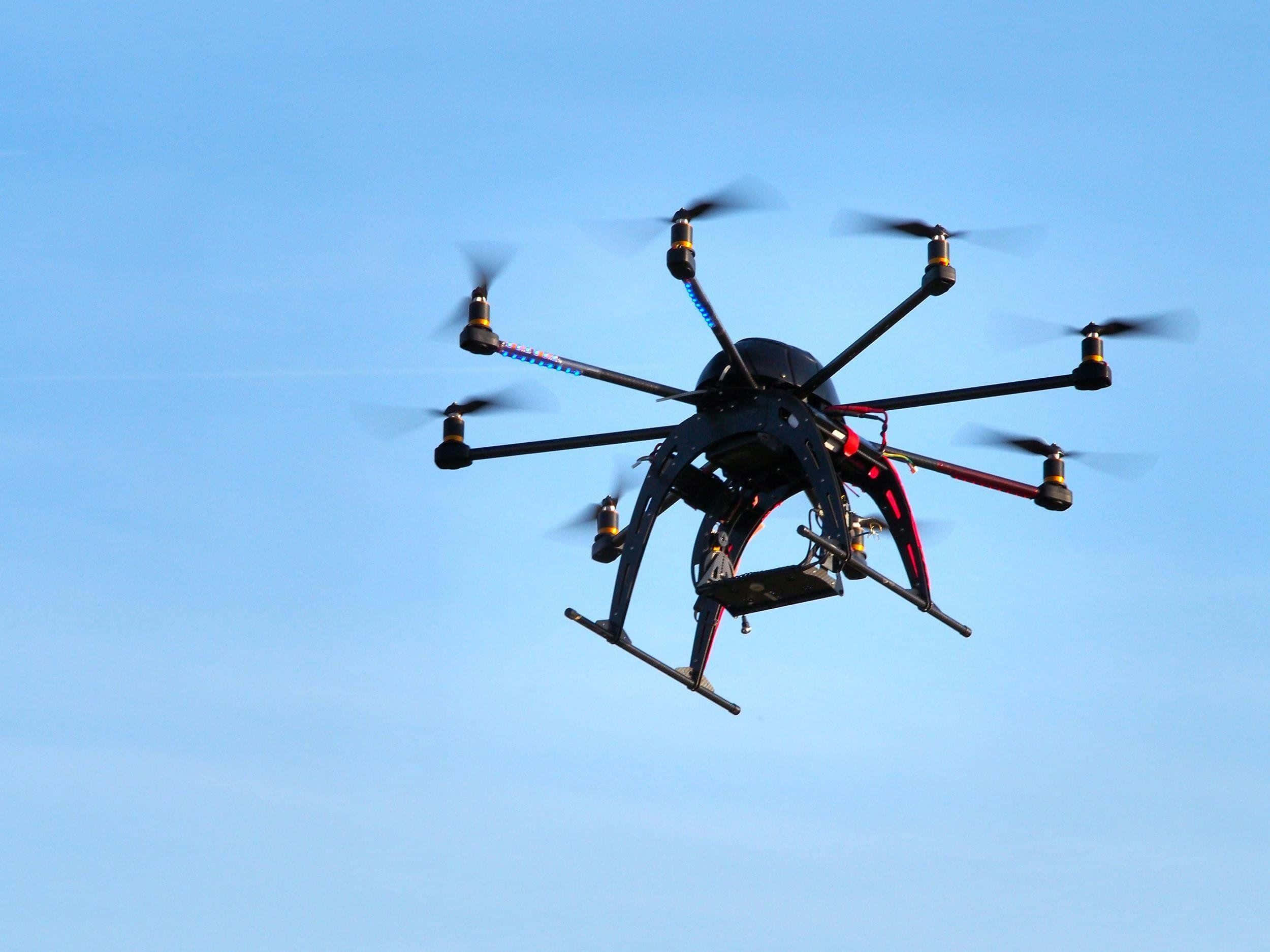 Drone Camera 桌布 壁紙 — 一架帶有紅色點綴的黑色六軸無人機在晴朗的藍天中盤旋，其旋翼因運動而模糊。