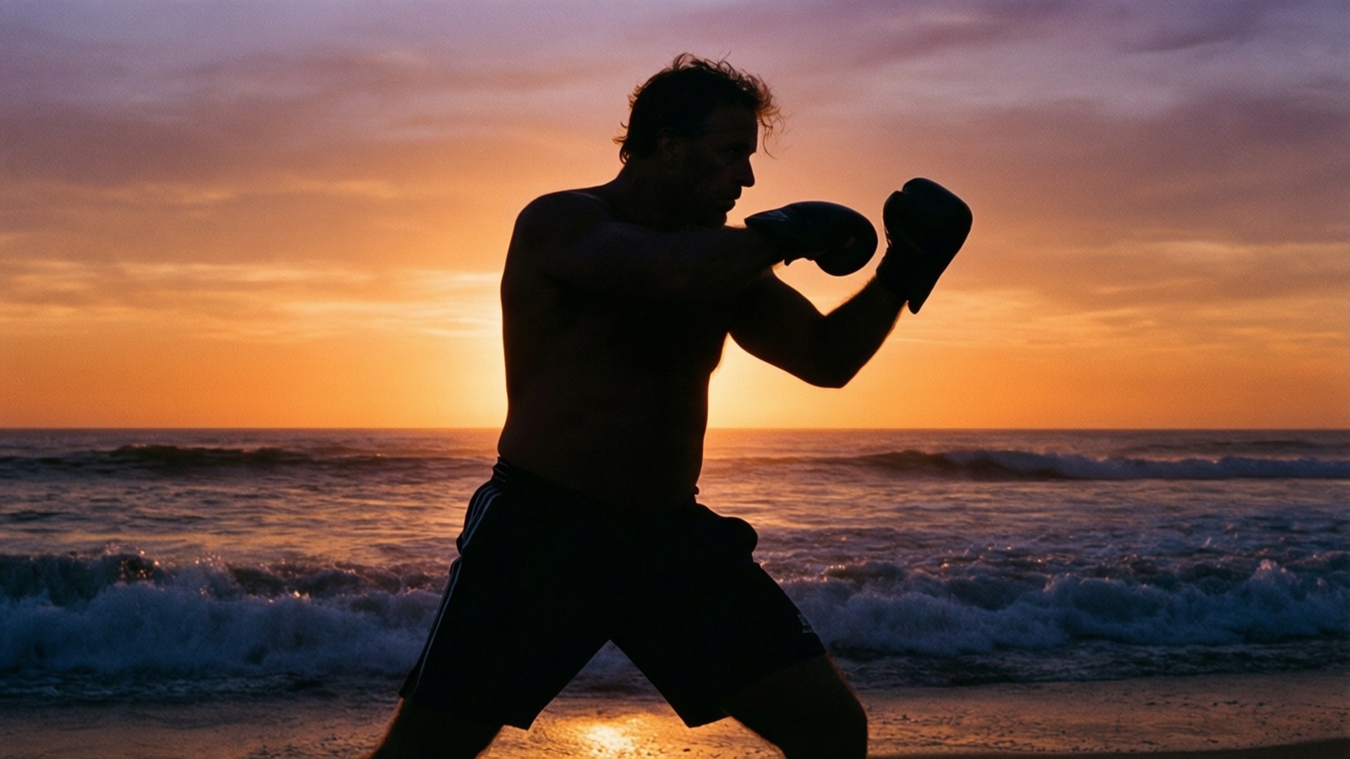 Sunrise Shadowboxing 壁紙 — 拳擊手的剪影在日落時分的海灘上訓練，身後波濤洶湧，天空火紅橙黃。