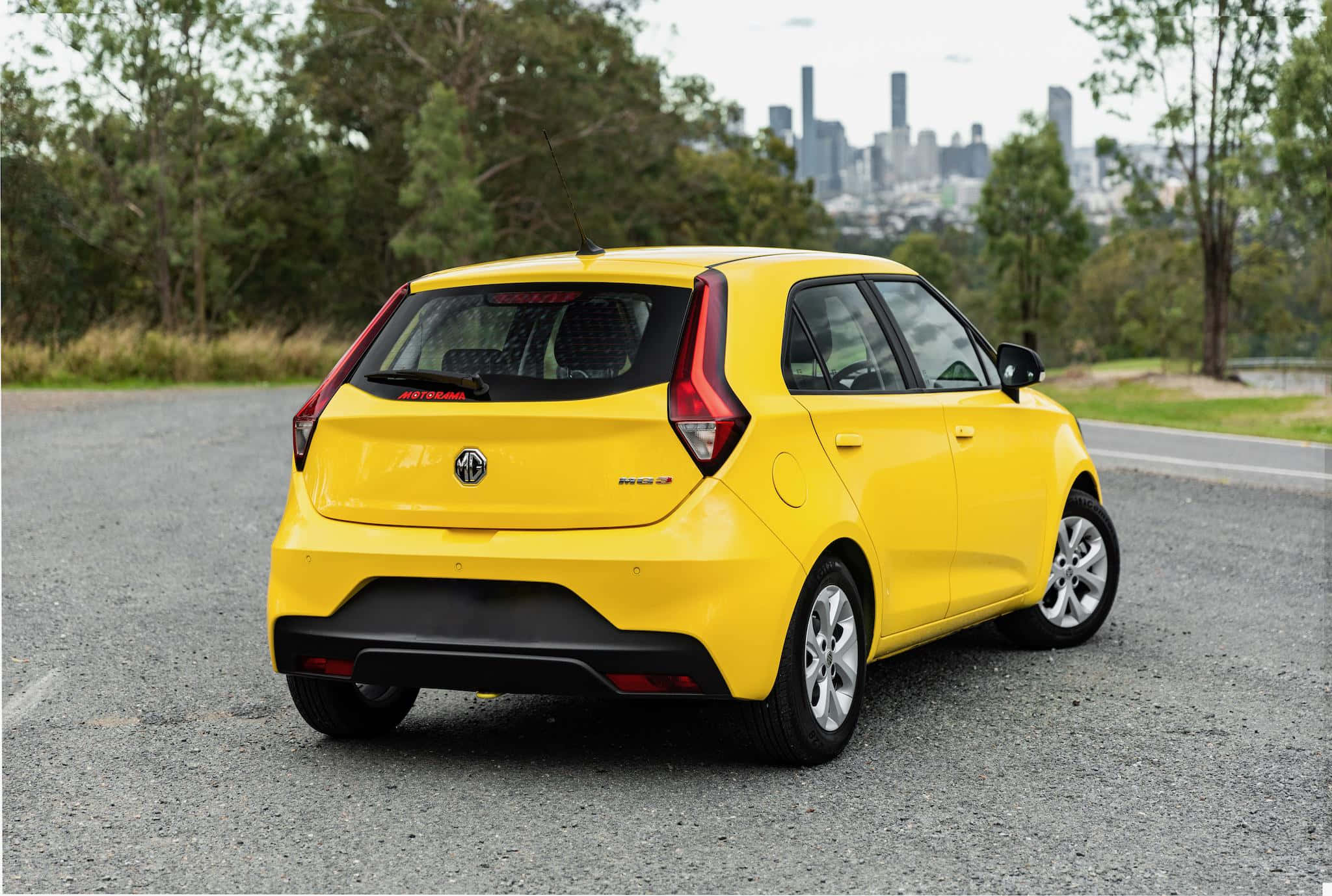Mg 3 桌布 壁紙 — 一輛亮黃色的MG汽車停在碎石上，背景是城市天際線和樹木。