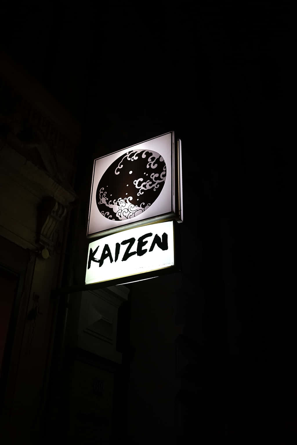  壁紙 — 一個明亮照明的「KAIZEN」霓虹燈牌，帶有波浪設計，在黑暗的夜晚街道上發光。