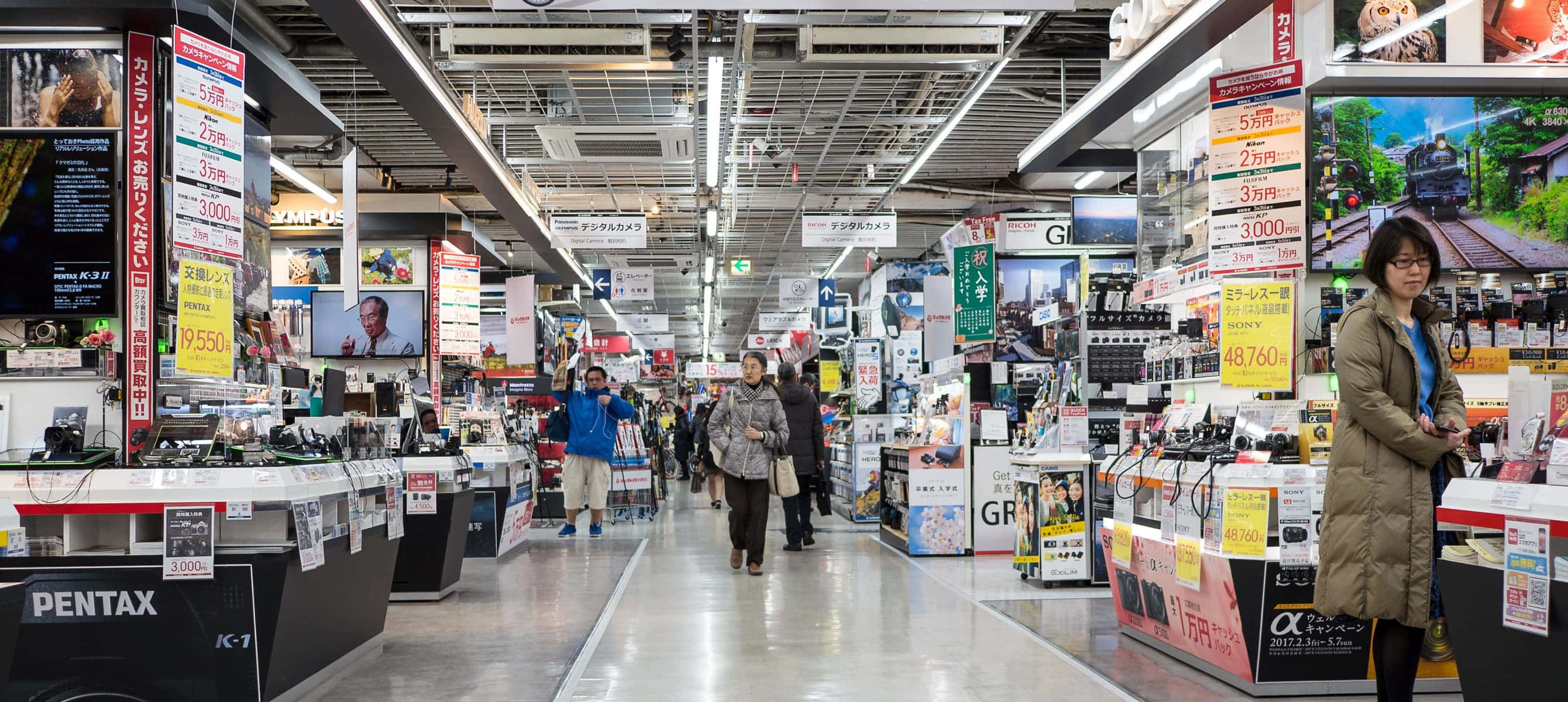 Store 桌布 壁紙 — 一條繁忙的電子產品商店通道，顧客在明亮的螢光燈下瀏覽相機和小工具。