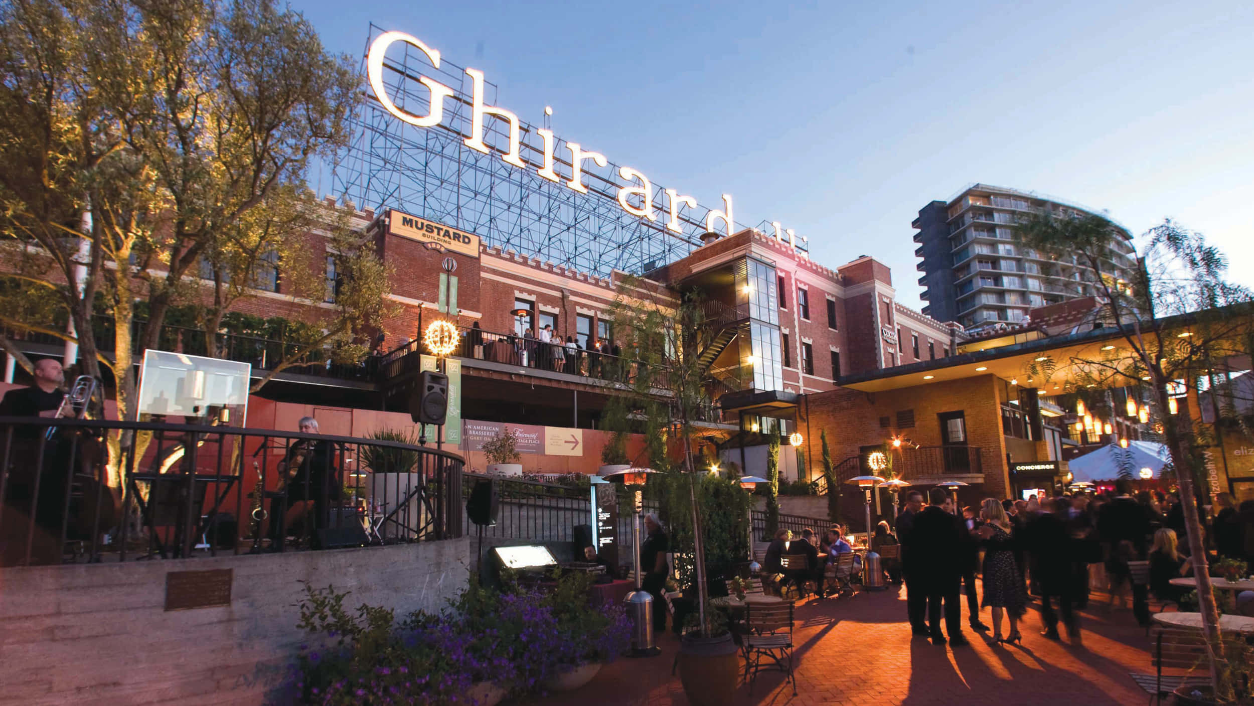 城市 桌布 壁紙 — Ghirardelli Square 一個熱鬧的戶外庭院，沐浴在溫暖的晚光中，人們在發光的串燈下用餐和散步。