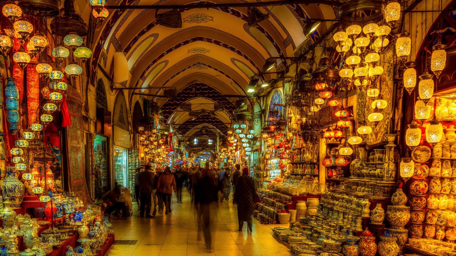 Grand Bazaar Colorful Chaos 壁紙 — 一條熱鬧的傳統市場走廊，由發光的燈籠照亮，兩旁擺滿色彩繽紛的陶瓷製品和紡織品。
