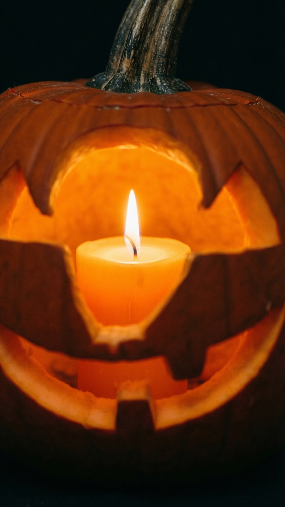 Candlelit Pumpkin 壁紙 — 一個雕刻南瓜，裡面有一根蠟燭，在黑色背景下溫暖地發光。