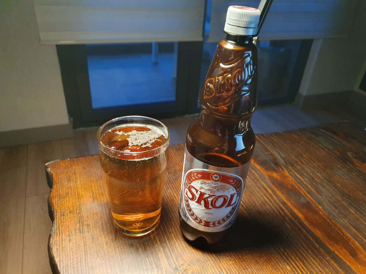  壁紙 — 一瓶冰鎮的Skol啤酒和酒杯放在靠近藍色玻璃窗的質樸木桌上。
