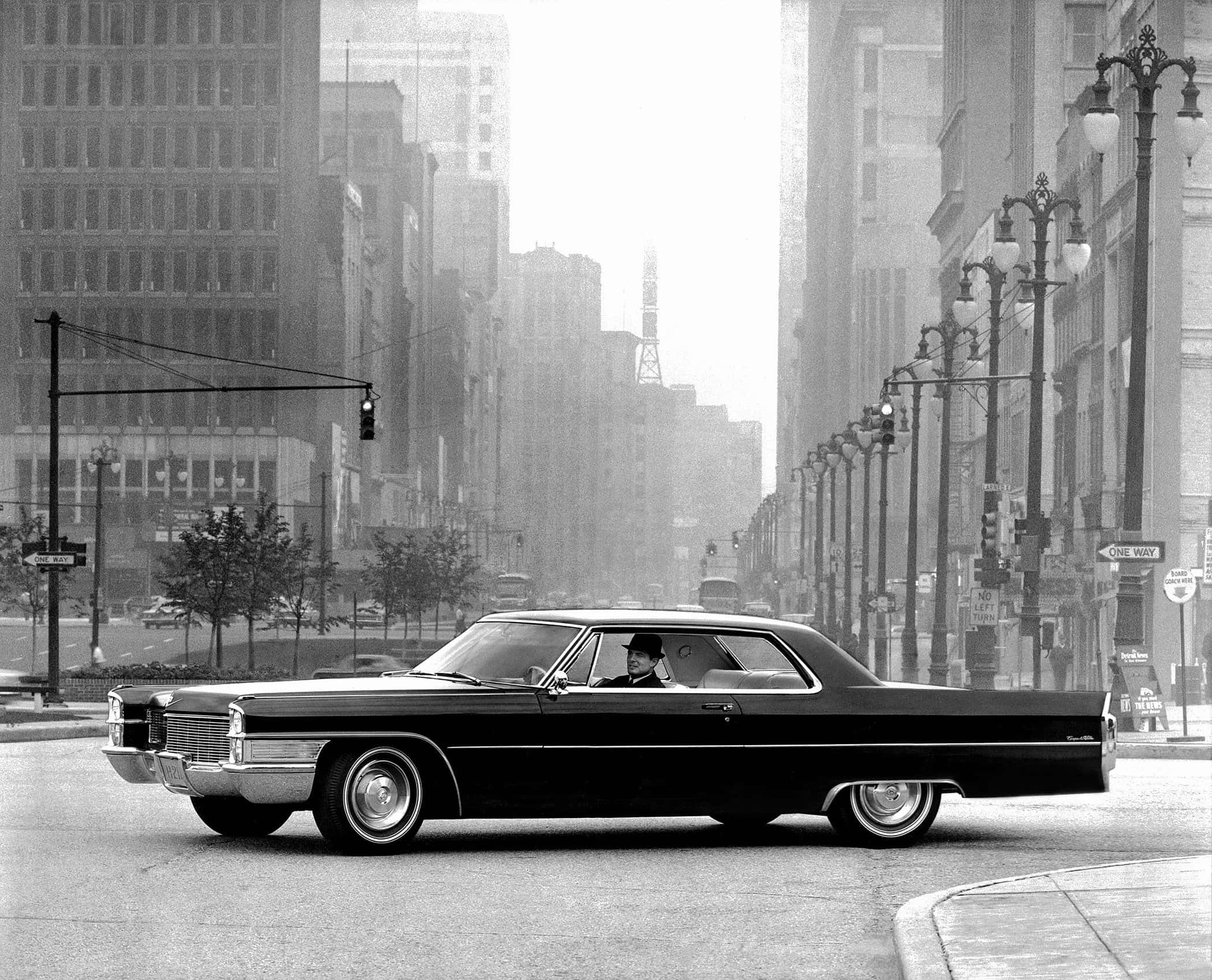 Cadillac Deville 桌布 壁紙 — 一輛經典黑色轎車在霧氣瀰漫的都市街道上行駛，兩旁是高樓大廈和復古路燈。