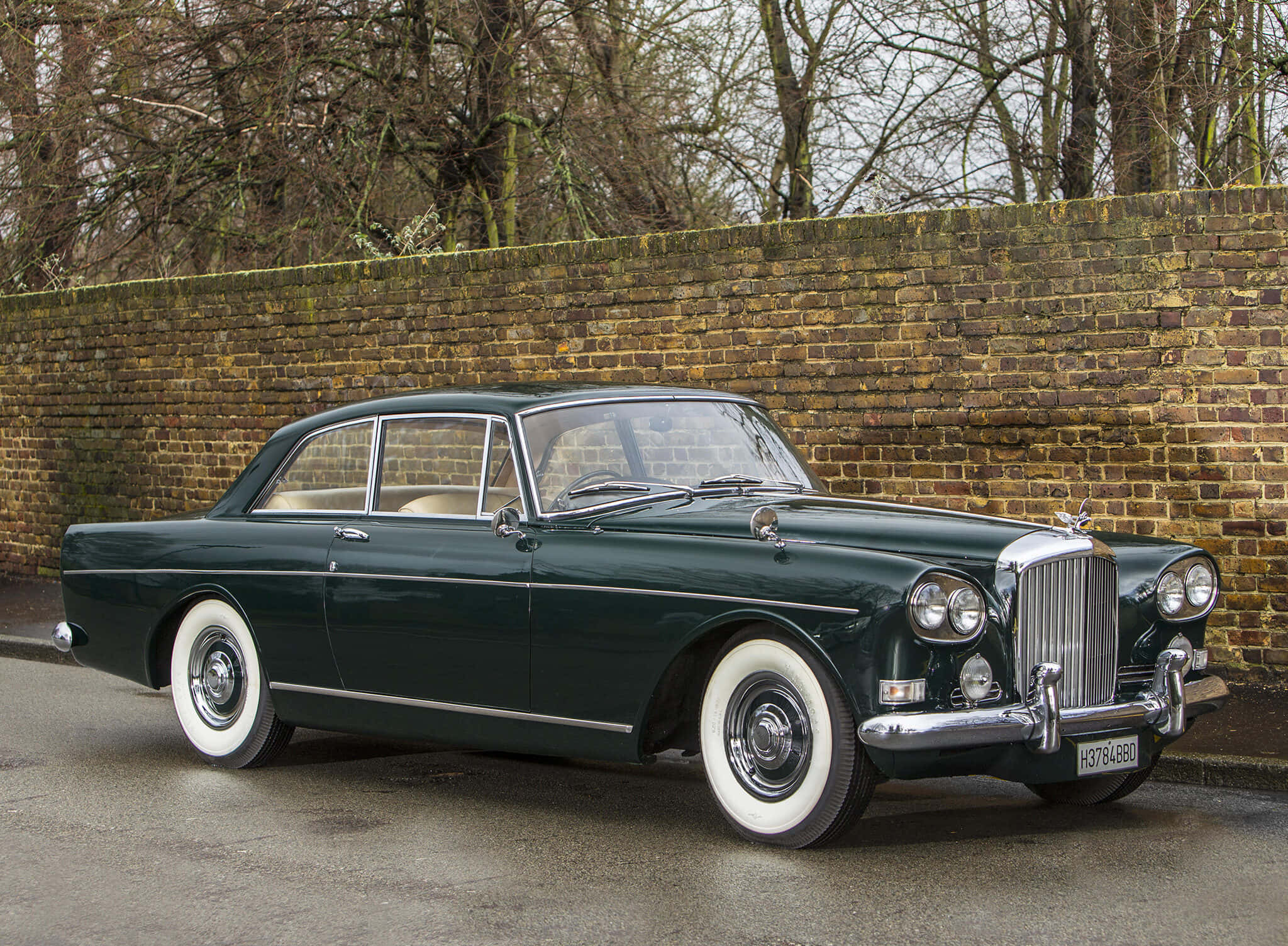 Bentley S3 桌布 壁紙 — 一輛經典綠色賓利配備白色側壁輪胎，停泊在裸露樹木下風化的磚牆旁。