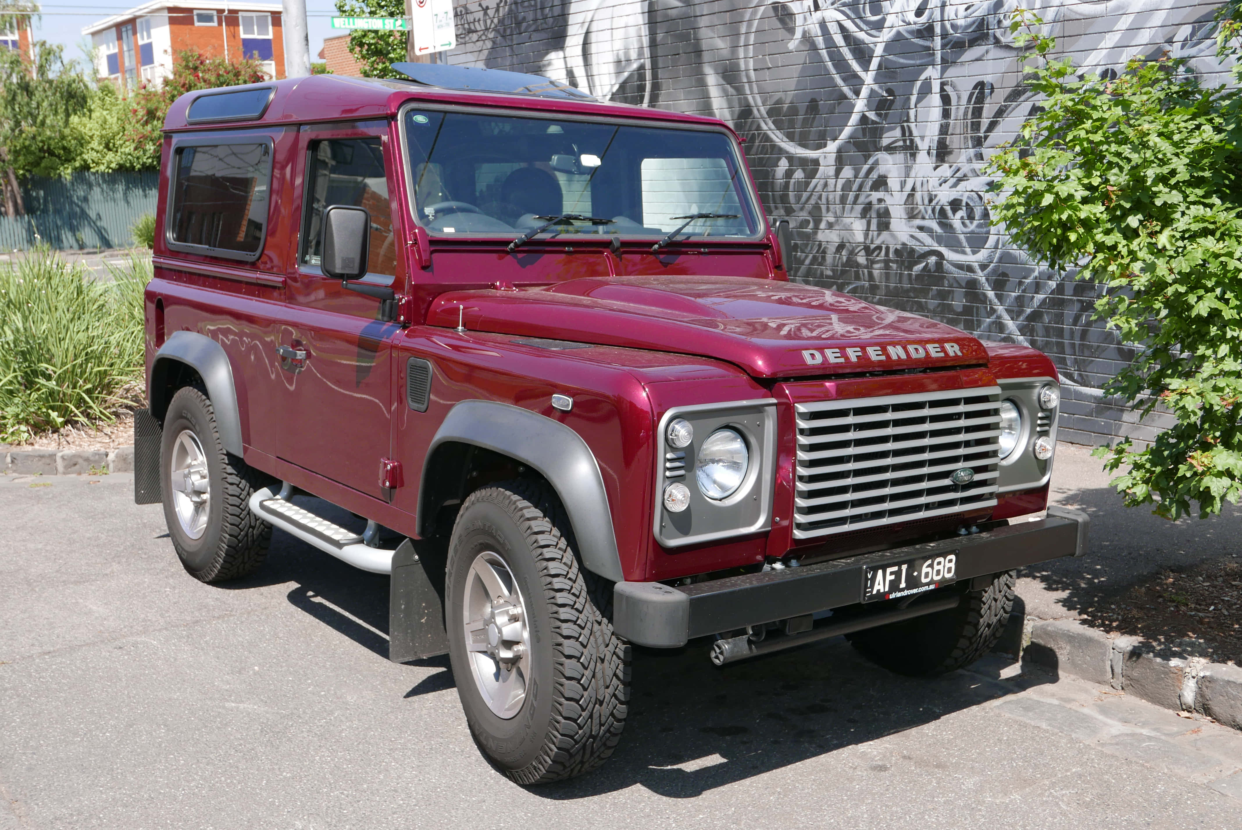 Land Rover Defender 桌布 壁紙 — 一輛經典的栗色Land Rover Defender停放在陽光明媚的街道上，旁邊是佈滿塗鴉的牆壁和綠植。