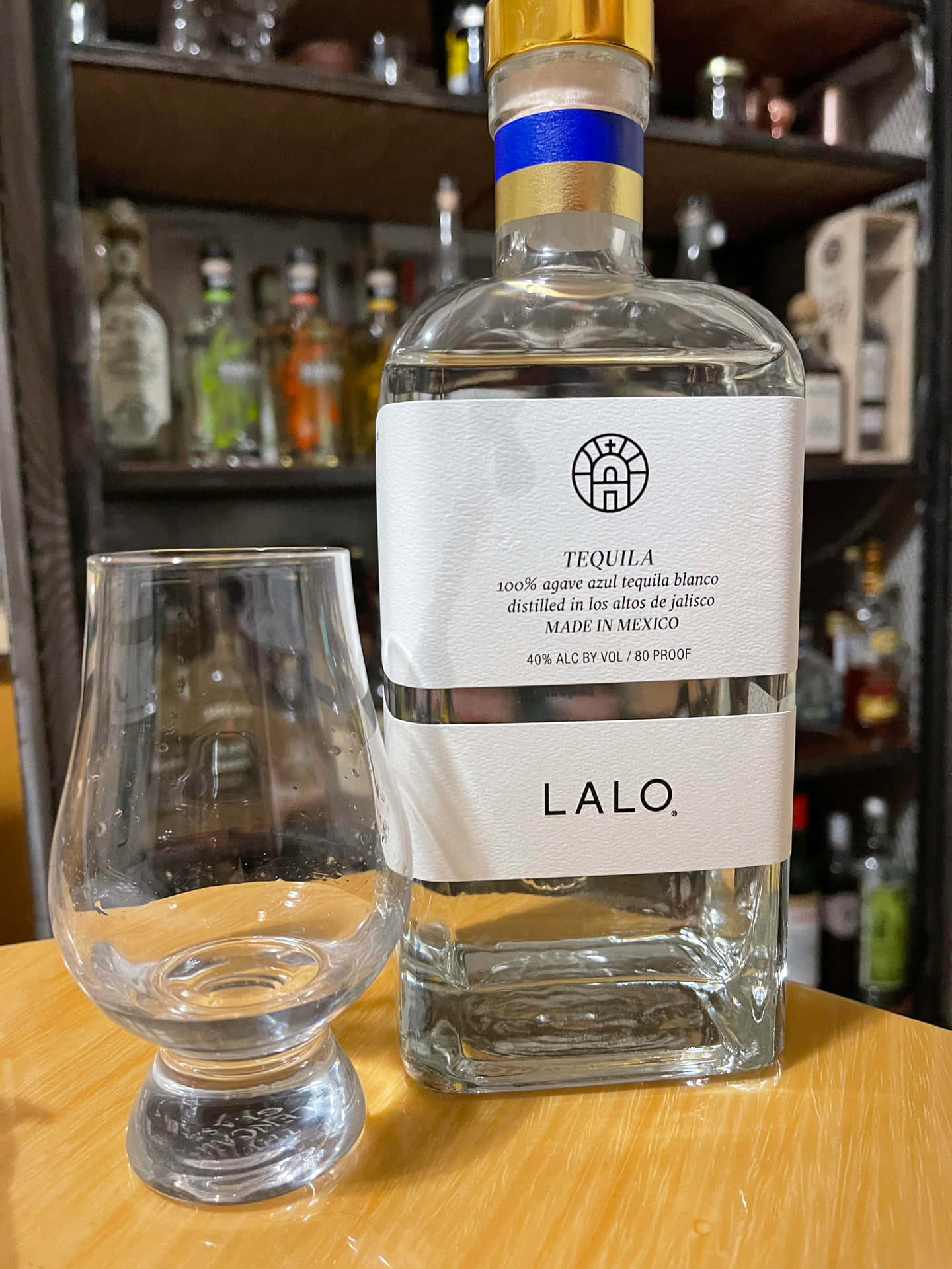  壁紙 — 一個透明的Lalo tequila blanco酒瓶放在一個空的品酒杯旁邊，背景是一個有酒架的木吧檯。