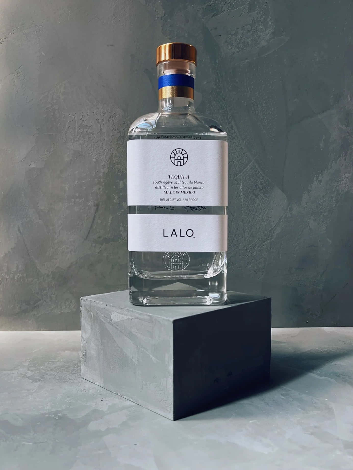  壁紙 — 一個透明的Lalo龍舌蘭酒瓶，金色瓶蓋，優雅地放置在灰色石座上，背景是帶有紋理的灰色牆壁。