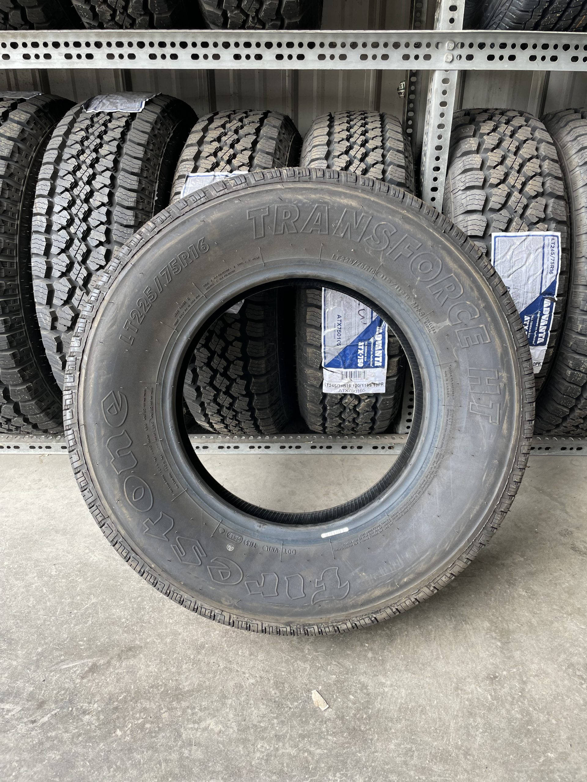 手機 桌布 壁紙 — Firestone TransForce HT 輪胎的特寫，放置在混凝土樓板上，後方金屬貨架上堆疊著其他輪胎。