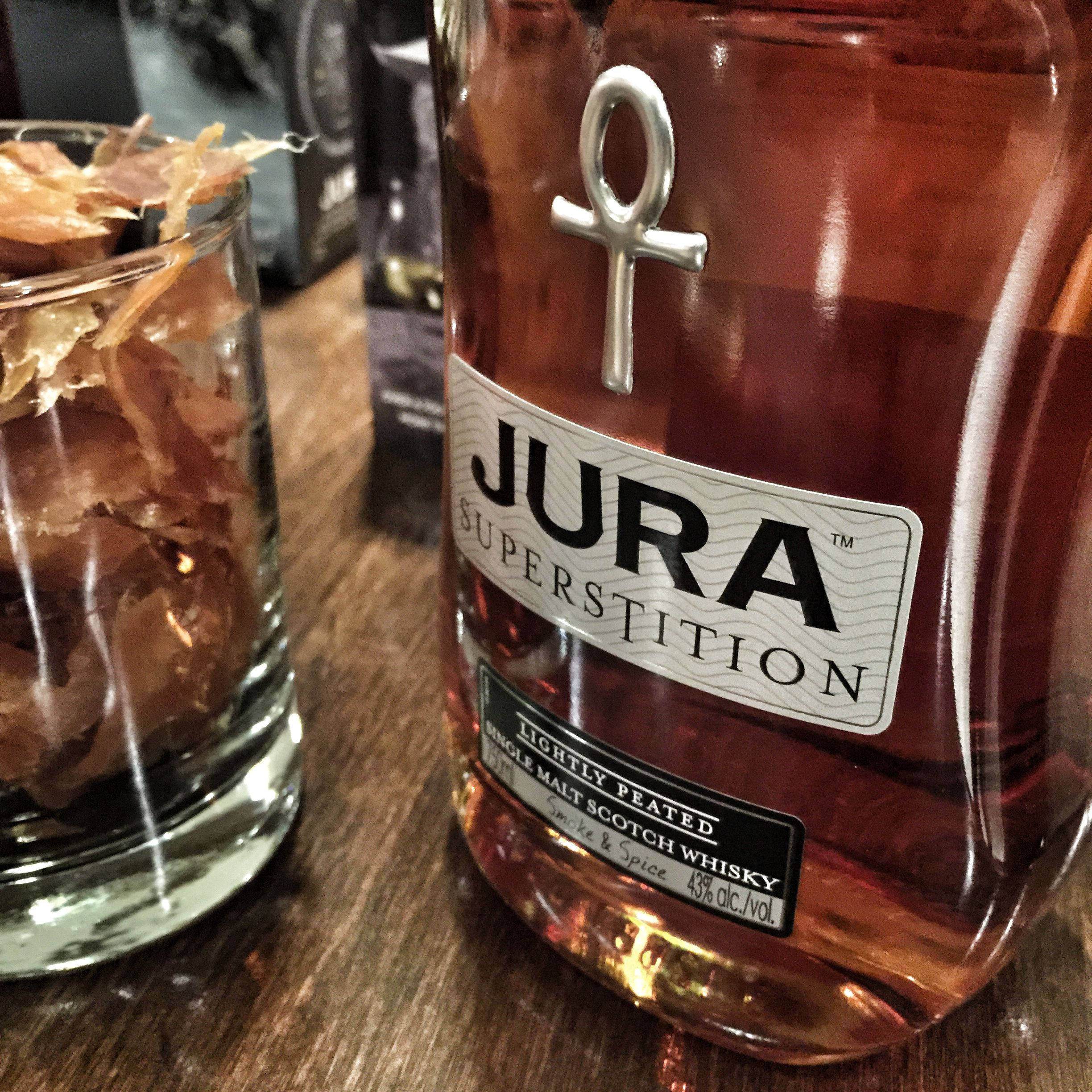 手機 桌布 壁紙 — Jura Superstition 威士忌酒瓶的特寫，帶有安卡標誌，旁邊是一個裝有乾木屑的玻璃杯，放在木桌上。