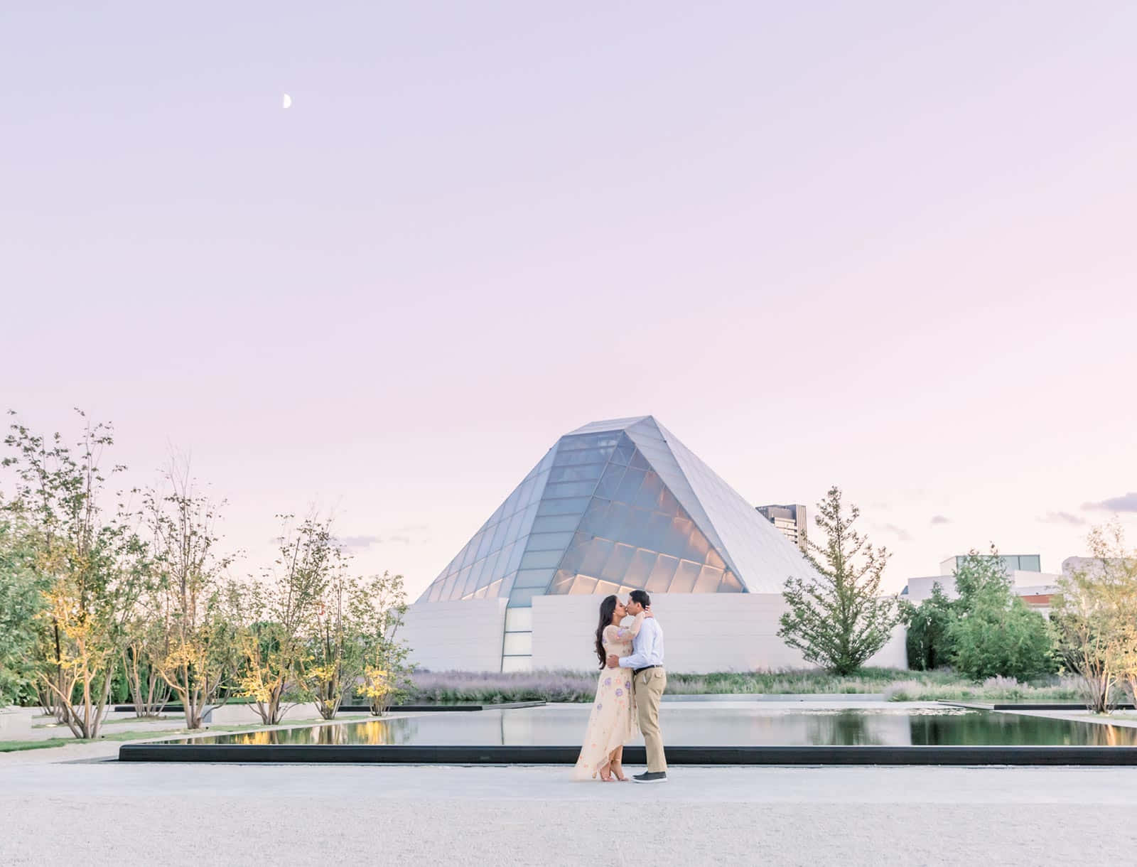 Aga Khan Museum 桌布 壁紙 — 一對情侶在黃昏時分，於玻璃金字塔建築前親吻，上方有一彎新月。
