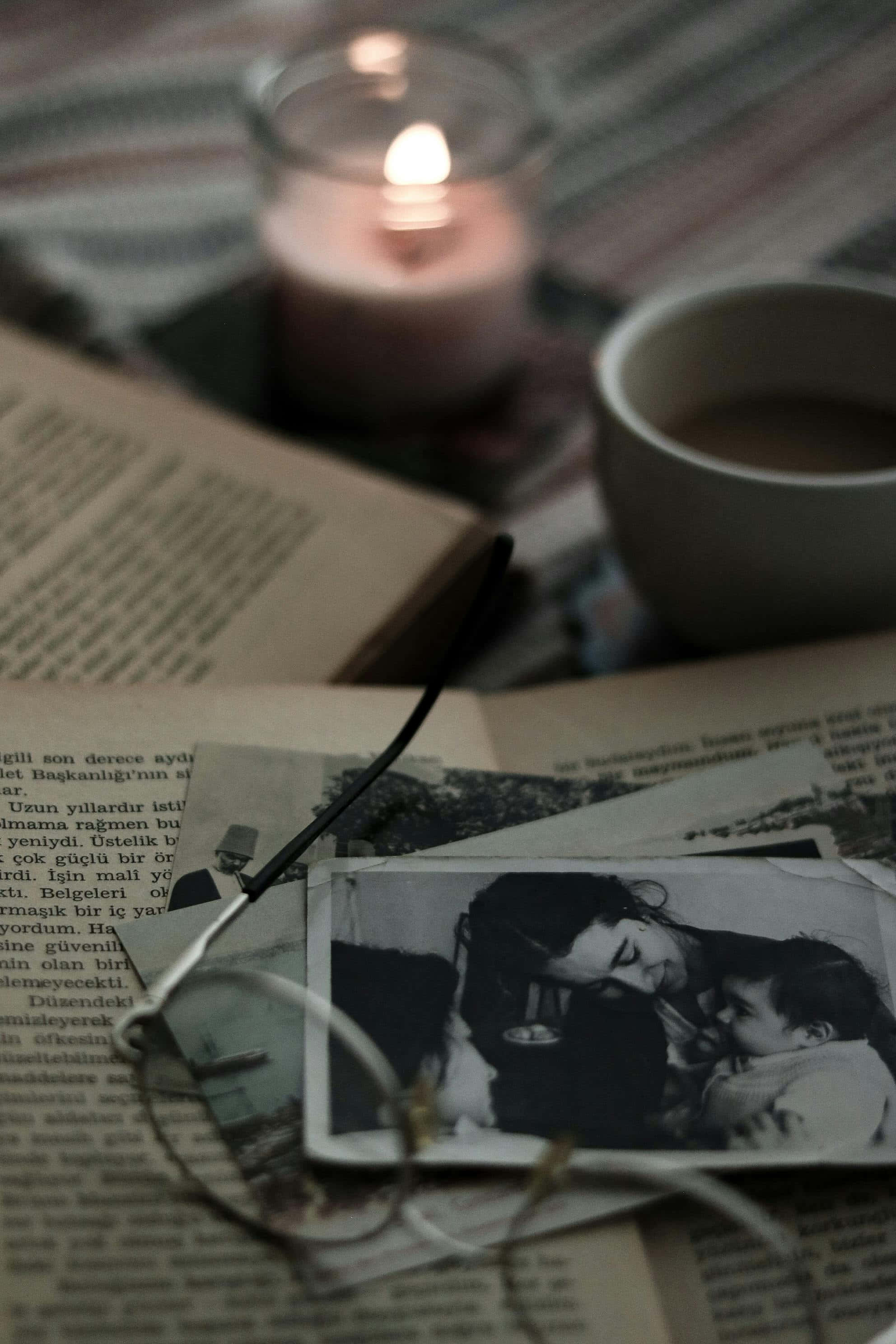 Dark Book Aesthetic 桌布 壁紙 — 一個舒適的夜晚場景，有一根點燃的蠟燭、一杯咖啡和一幅復古照片放在一本打開的書上，附近有眼鏡。