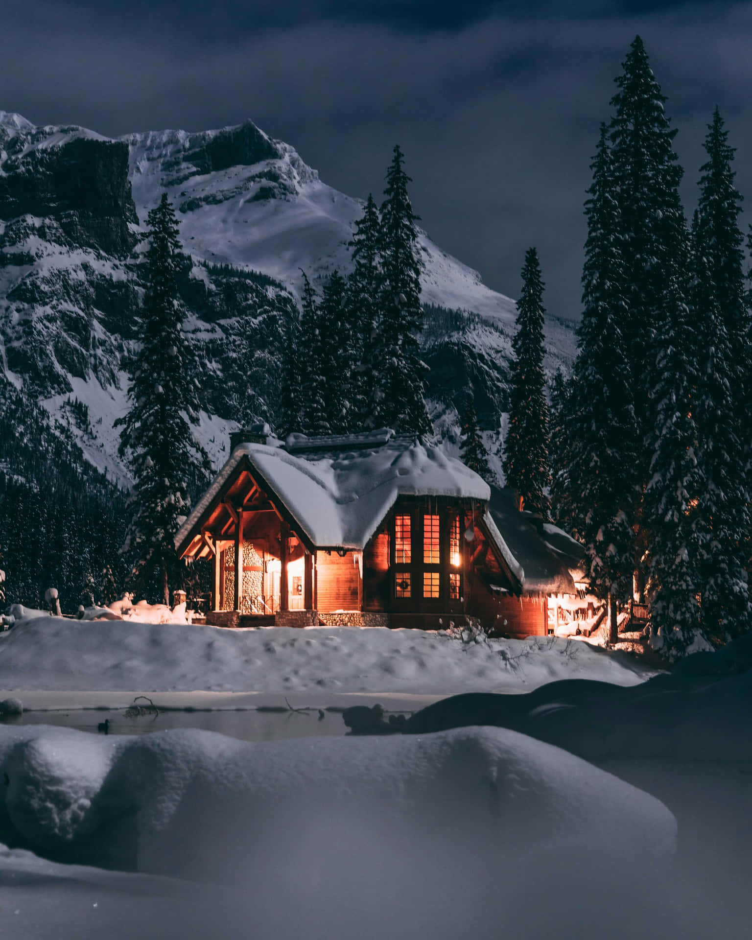  壁紙 — 一間舒適的雪覆蓋小屋在黑暗的山景背景下，在雪夜天空下溫暖地發光。