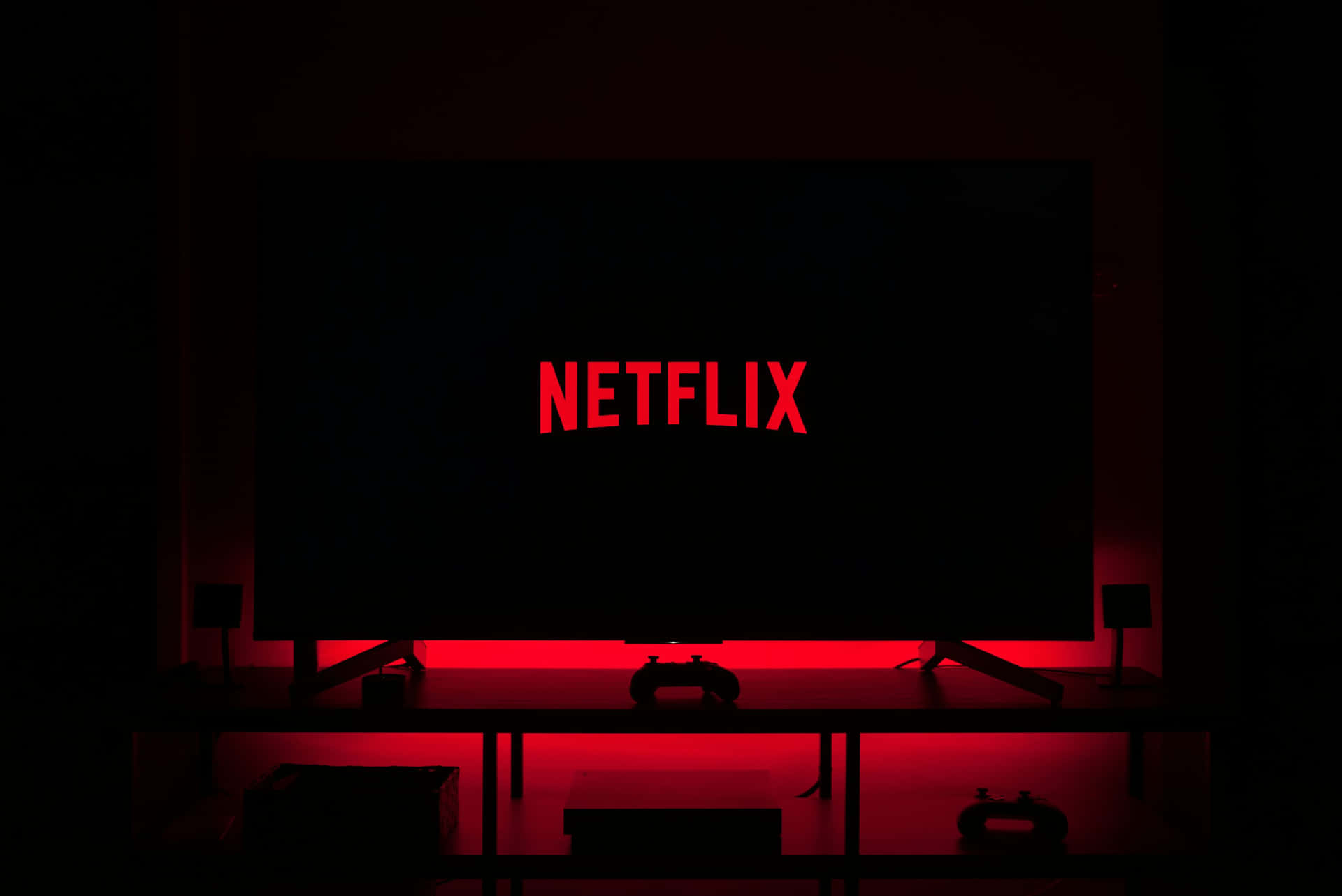  壁紙 — 一個黑暗的房間，被電視螢幕上發光的紅色Netflix標誌和下方的遊戲控制器照亮。