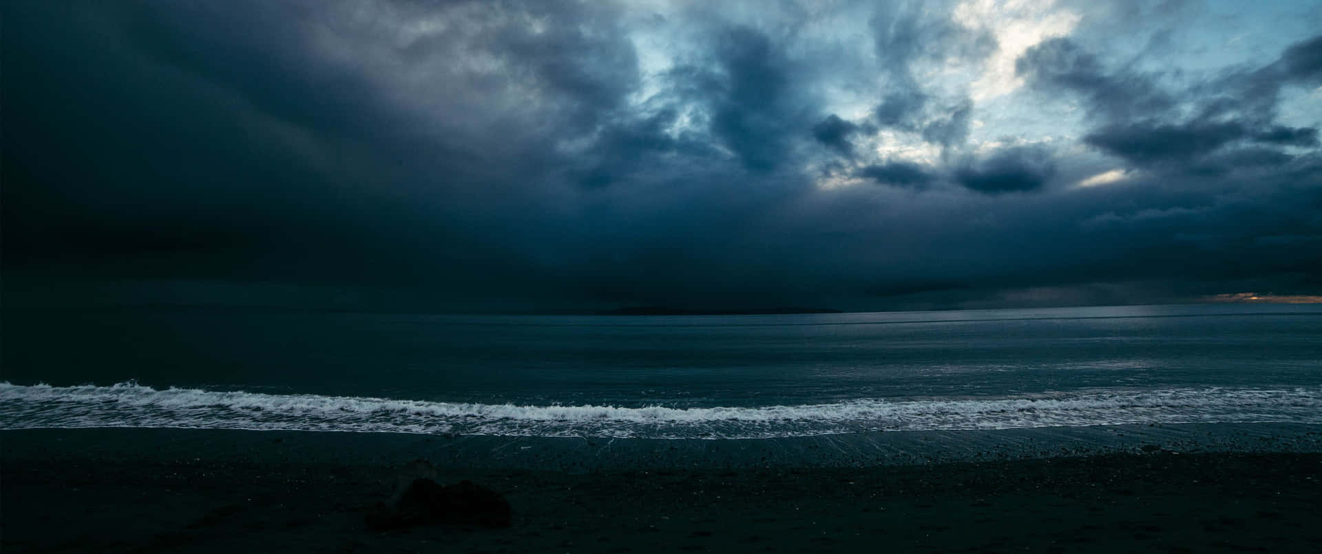 黑暗的海浪在黃昏時分厚重的雲層下拍打著暴風雨的海灘。 — Oled 壁紙