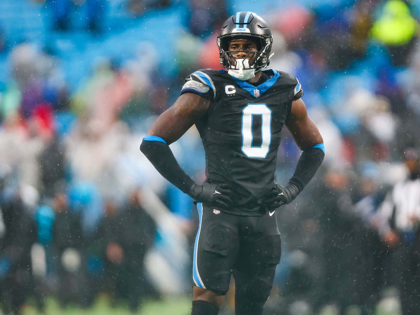  壁紙 — 一位堅定的NFL球員身穿黑藍色制服，雙手叉腰站在下雨的足球場上。