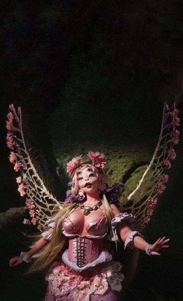  壁紙 — 一個像娃娃的仙女，擁有粉紅色花卉翅膀和緊身胸衣，站在黑暗的森林中，被柔和的光線照亮。