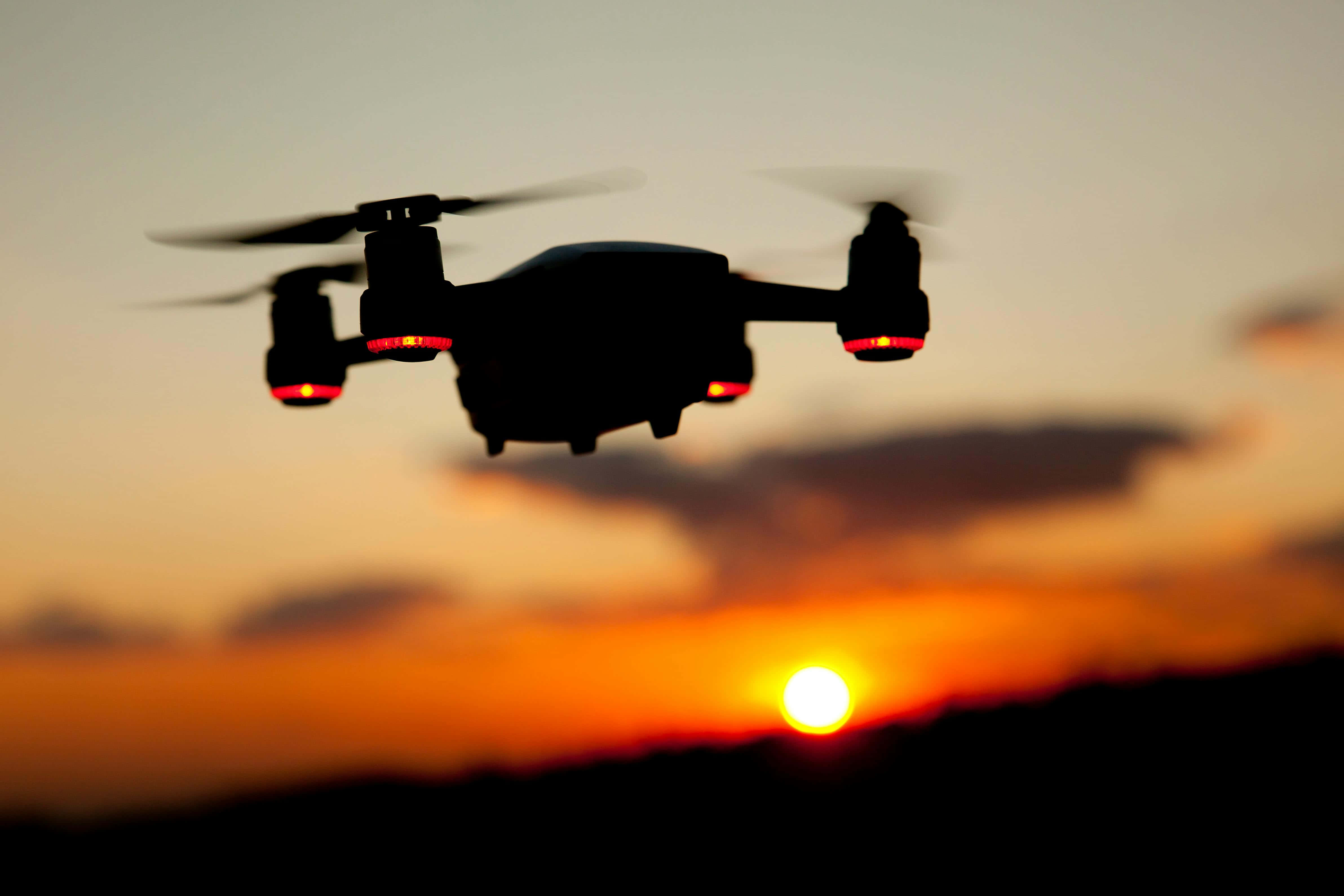 Drone Camera 桌布 壁紙 — 一架無人機在鮮豔的日落天空襯托下形成剪影，帶有發光的紅燈和模糊的螺旋槳。