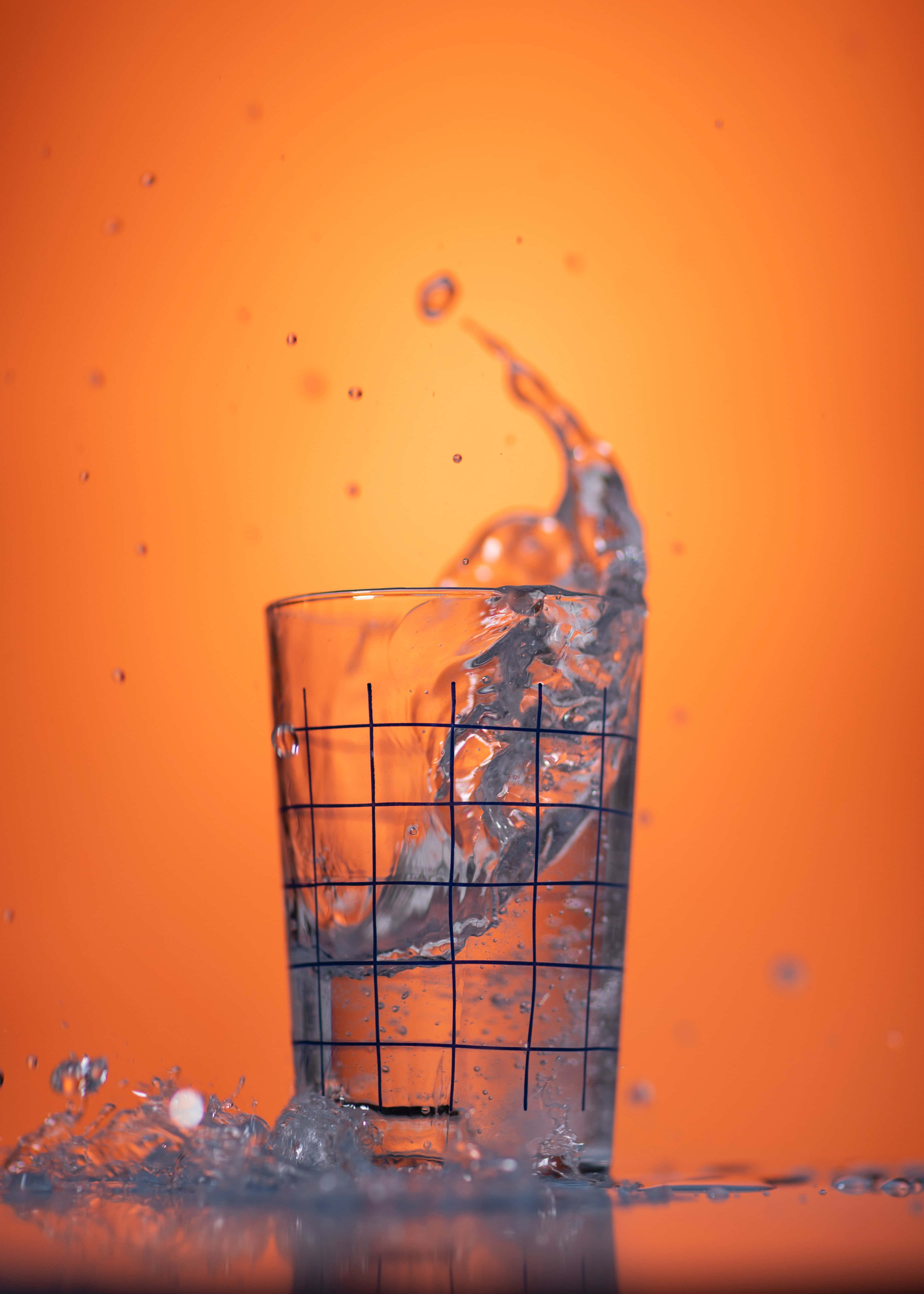 水的極樂：滋養生命之源。 壁紙 — 一個動態的水花從玻璃杯中噴出，背景是充滿活力的橙色。