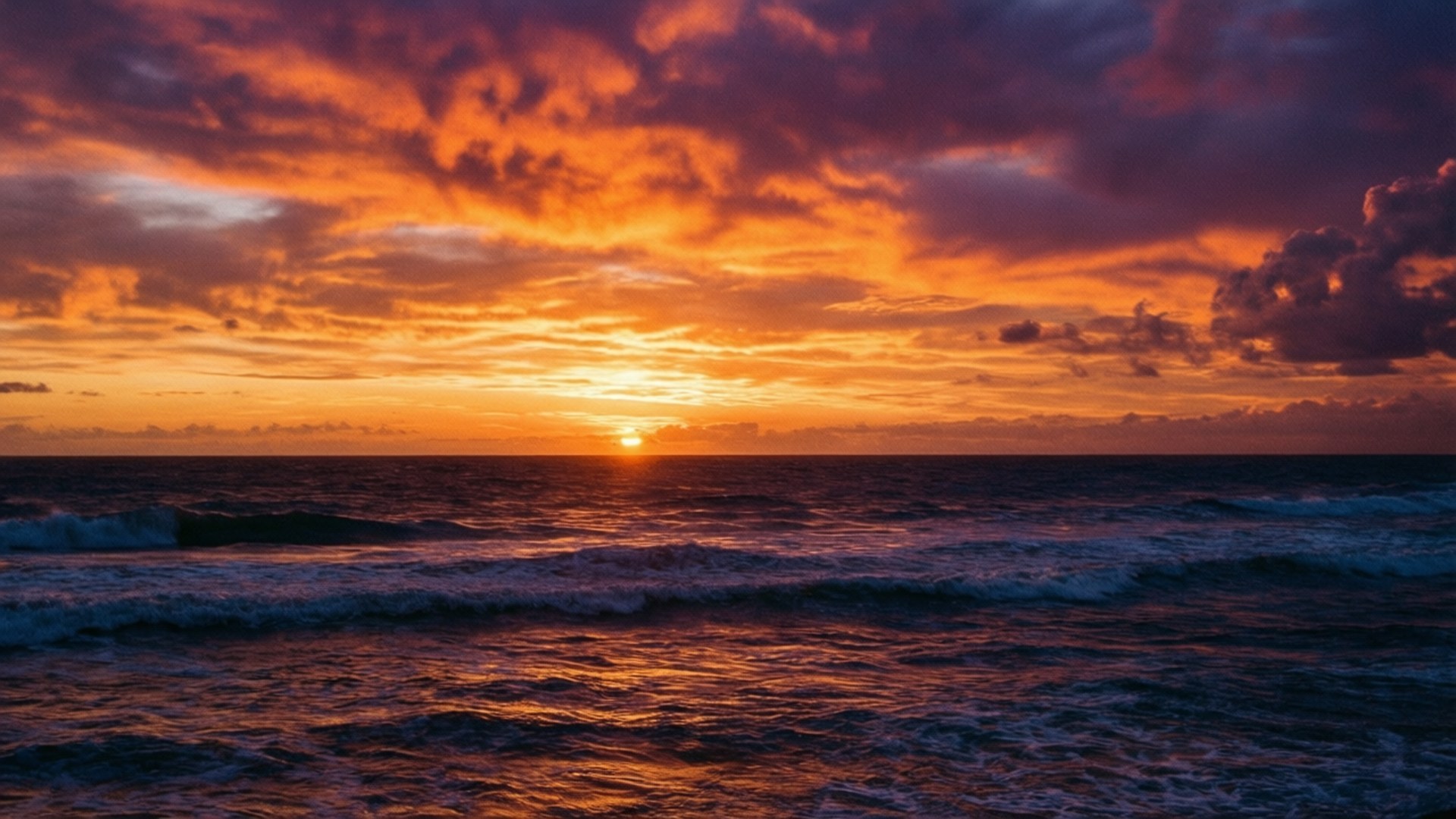 Fiery Ocean Sunset 壁紙 — 熾熱的夕陽將海洋染成橙色和紫色，海浪在戲劇性的多雲天空下拍打。