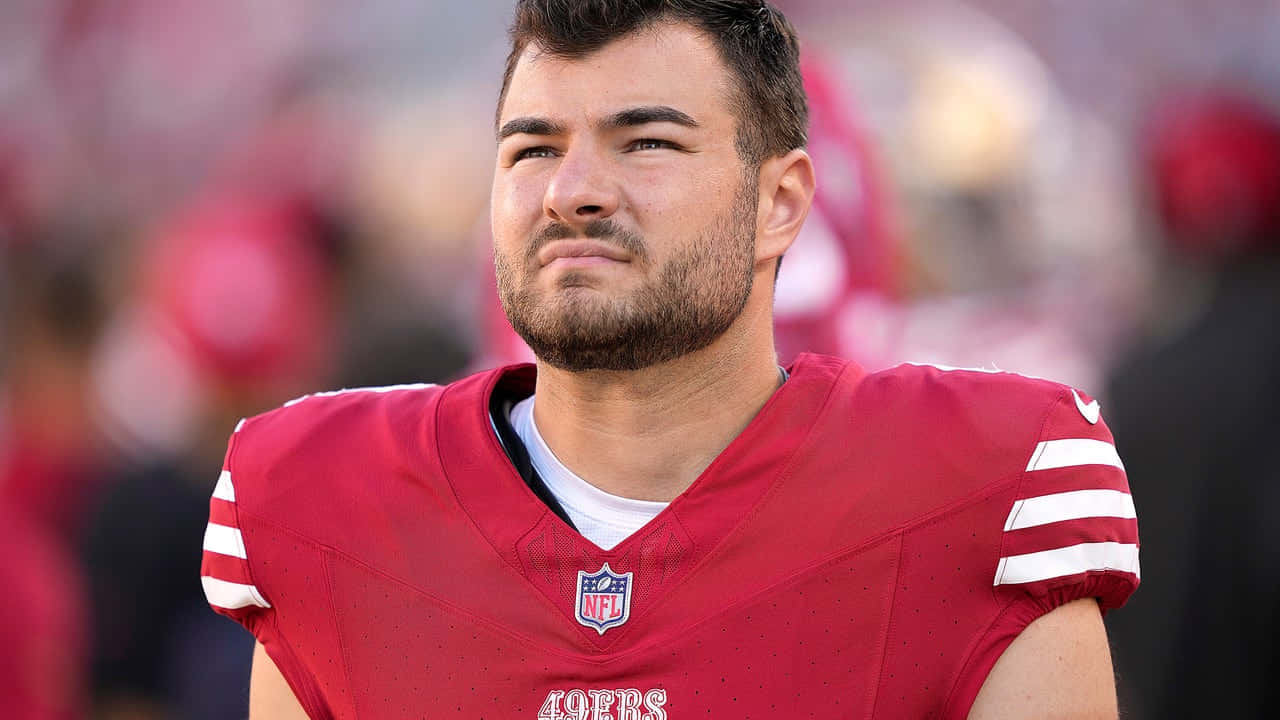  壁紙 — 一位專注的NFL美式足球員身穿紅色49ers球衣，帶著堅定的表情站在球場上。