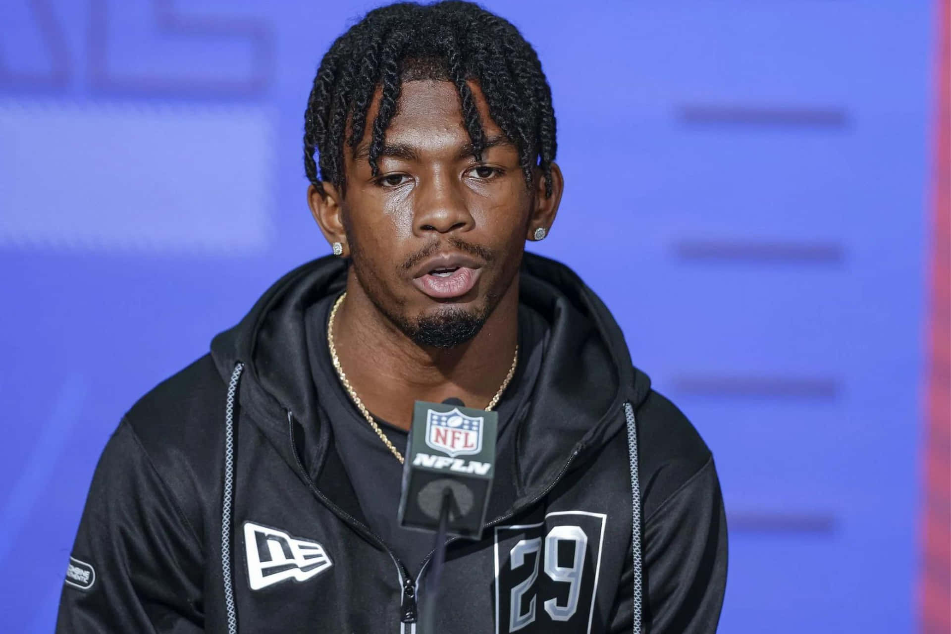  壁紙 — 一位專注的NFL球員身穿黑色連帽衫，號碼29，在藍色背景前對著NFL麥克風說話。