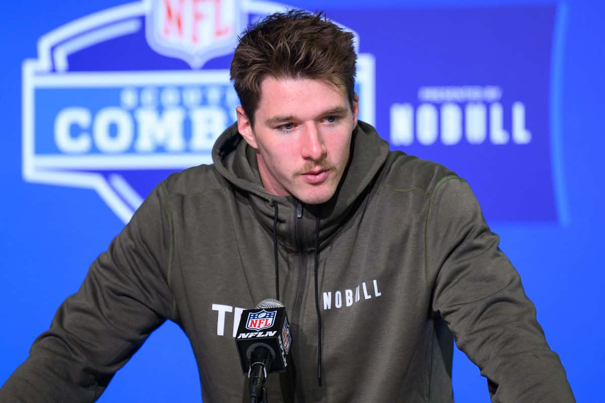  壁紙 — 一位專注的NFL球員穿著綠色連帽衫，在藍色背景和麥克風的新聞發布會上發言。