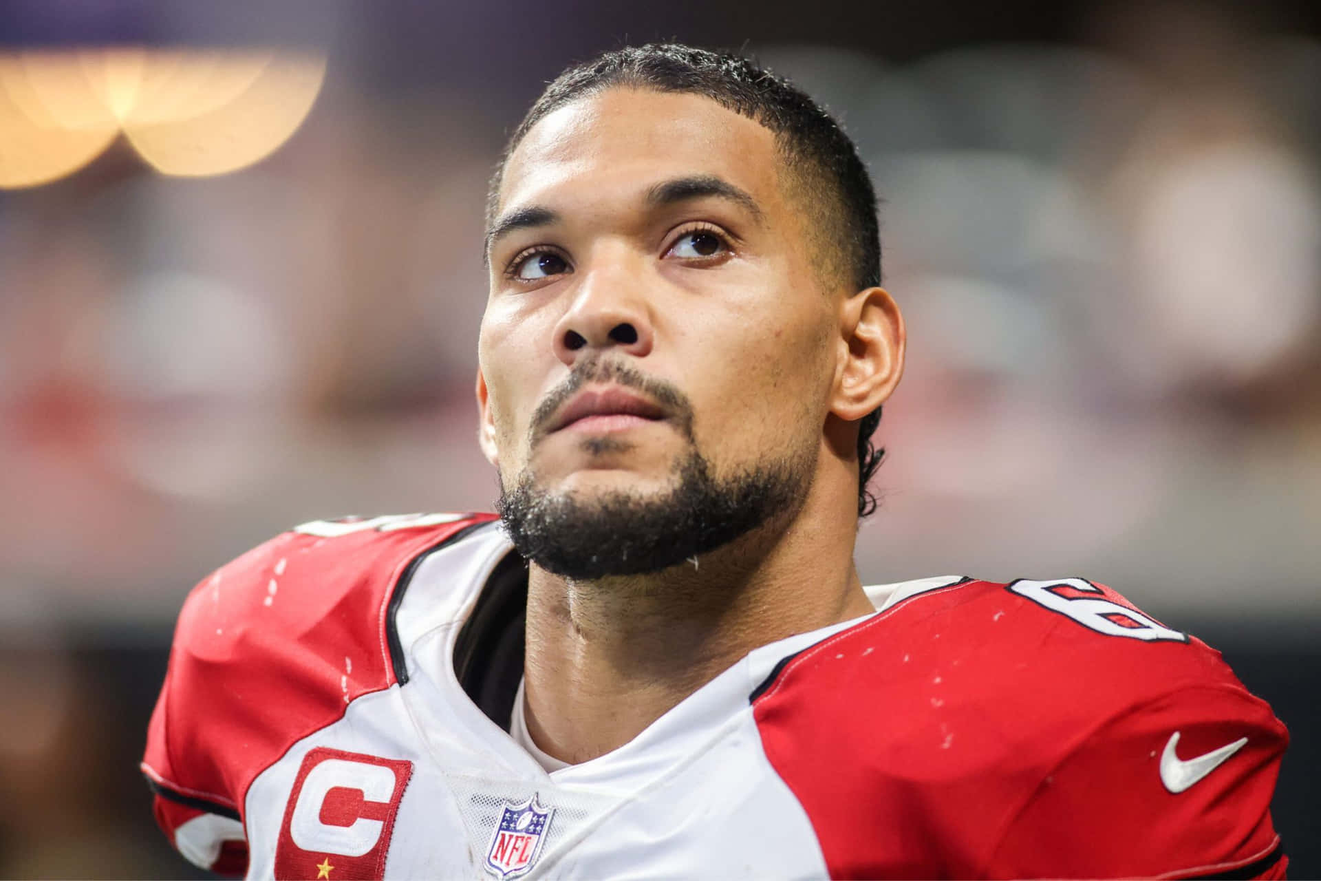  壁紙 — 一位專注的NFL球員身穿紅白球衣，帶有隊長'C'標誌，在球場燈光下向上凝視。