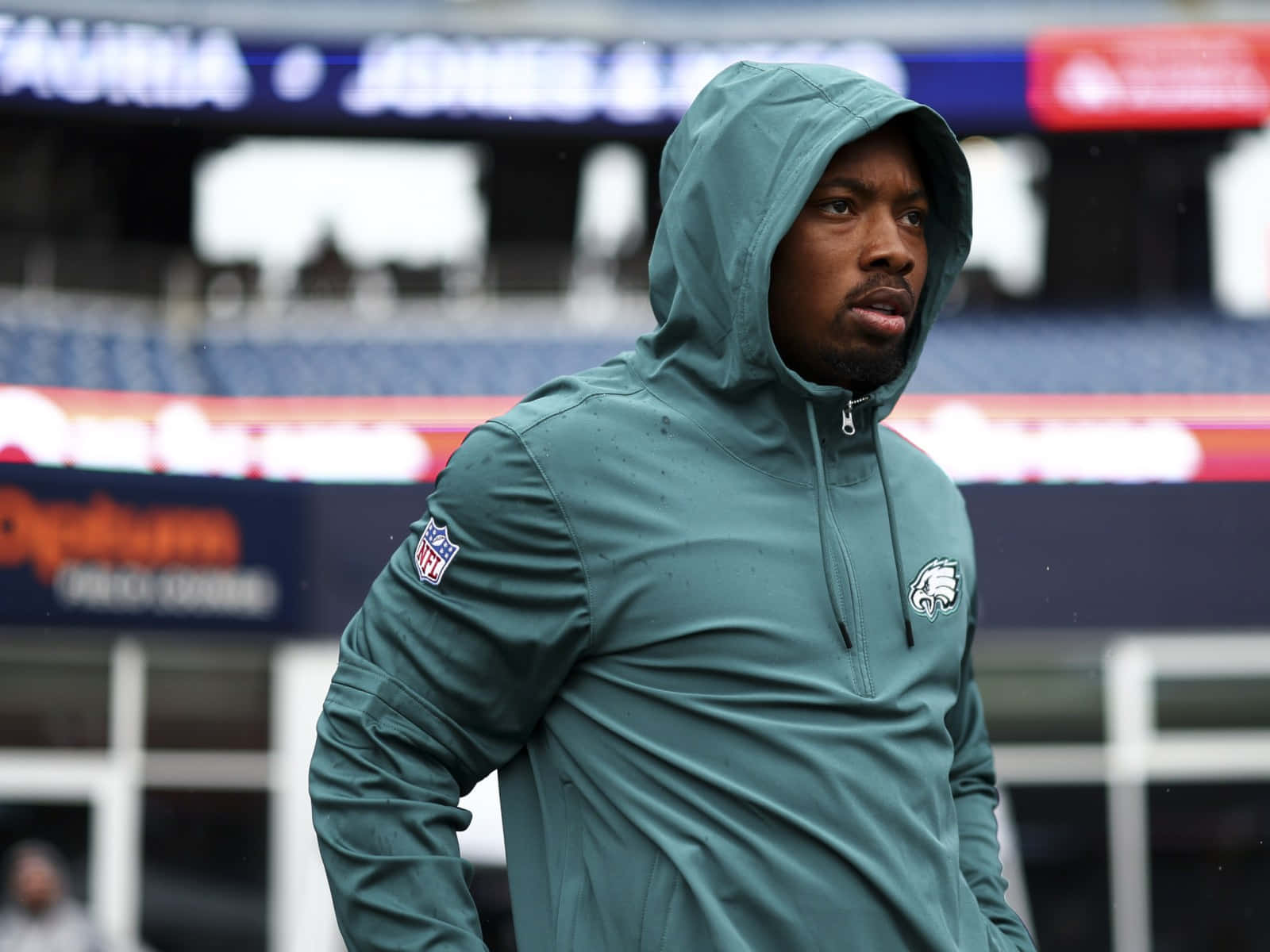  壁紙 — 一位專注的NFL球員穿著青綠色連帽衫，站在下雨的體育場球場上，身後是模糊的看台。