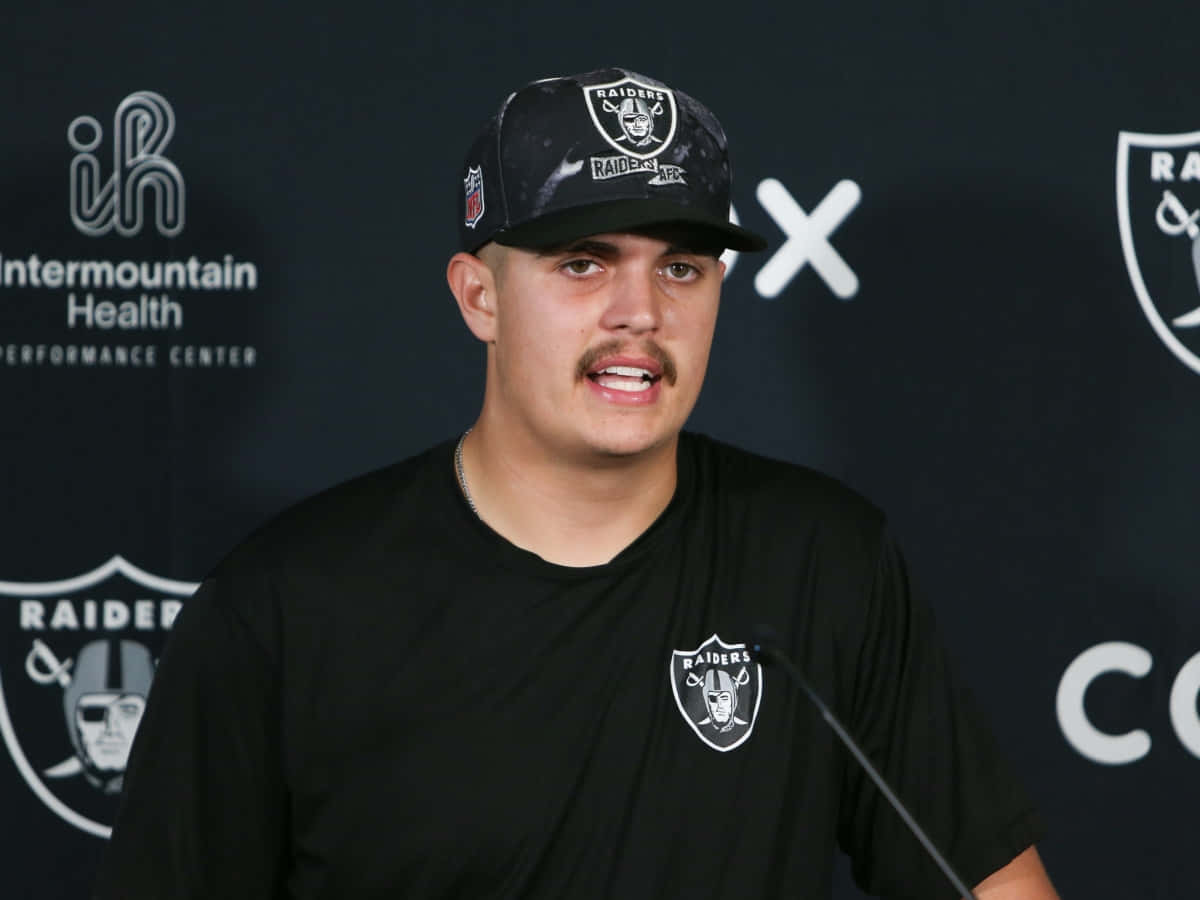 壁紙 — 一名戴著Raiders帽子和穿著Raiders襯衫的足球運動員在記者會上發言，背景為深色並帶有球隊標誌。