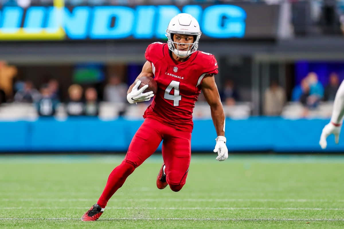  壁紙 — 一名身穿紅色 Cardinals 裝備的美式足球員在日間比賽中，抱著球衝過綠色球場。