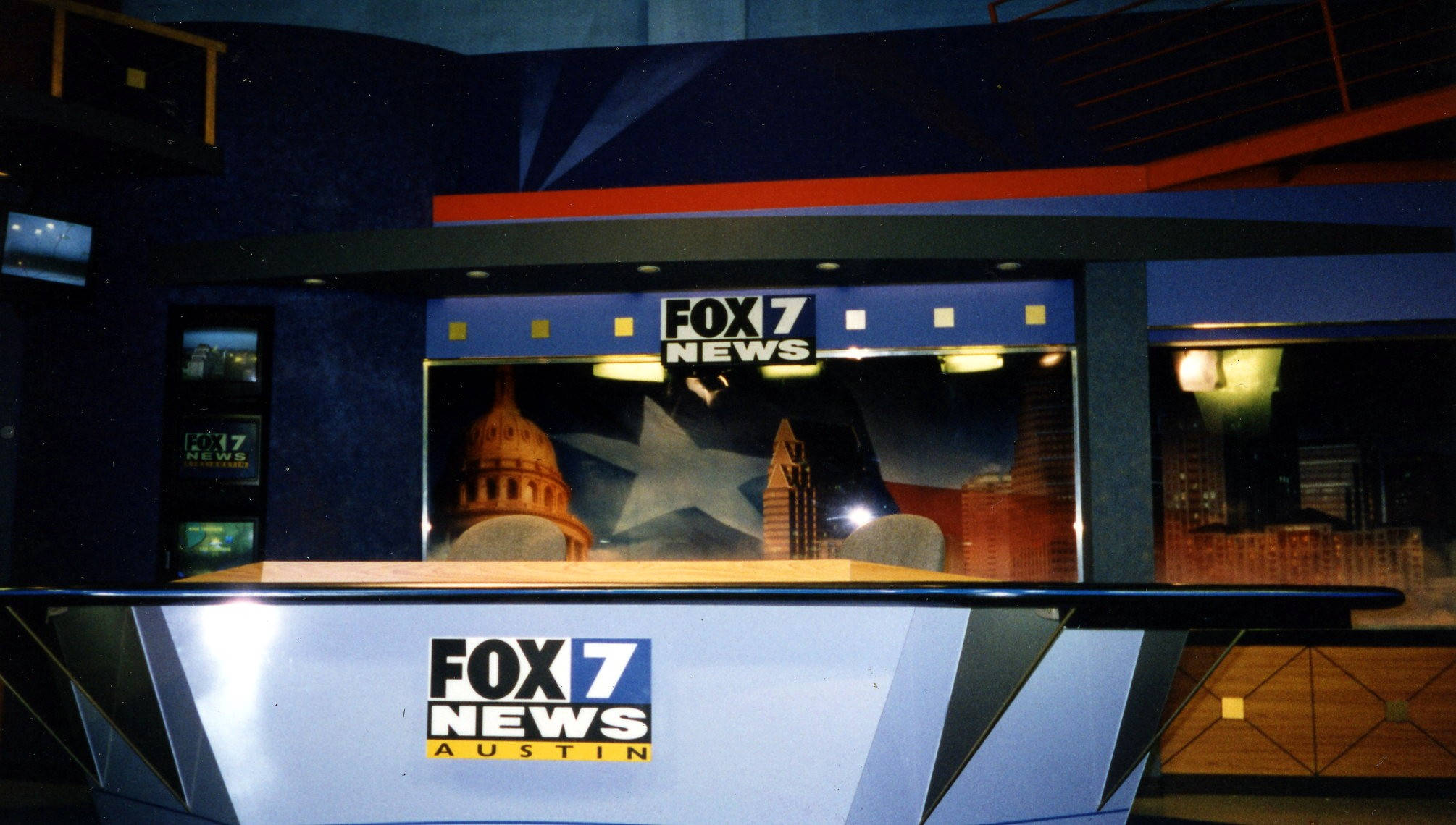 News 桌布 壁紙 — Fox 7 News Austin 的攝影棚佈景，設有藍色幾何牆壁，背景顯示夜間的德州州議會大廈和天際線。