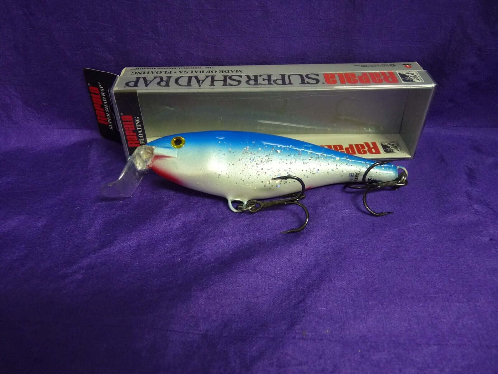  壁紙 — 一個帶有藍銀色和紅色塗漆的Rapala Super Shad Rap釣魚假餌，旁邊放著它的盒子，背景是紫色布料。