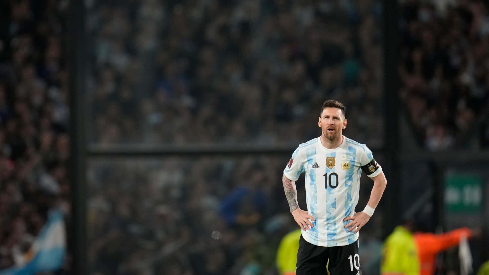 Messi 2022 桌布 壁紙 — 阿根廷足球巨星梅西身穿藍白條紋球衣，在明亮的燈光下雙手叉腰站在球場上。