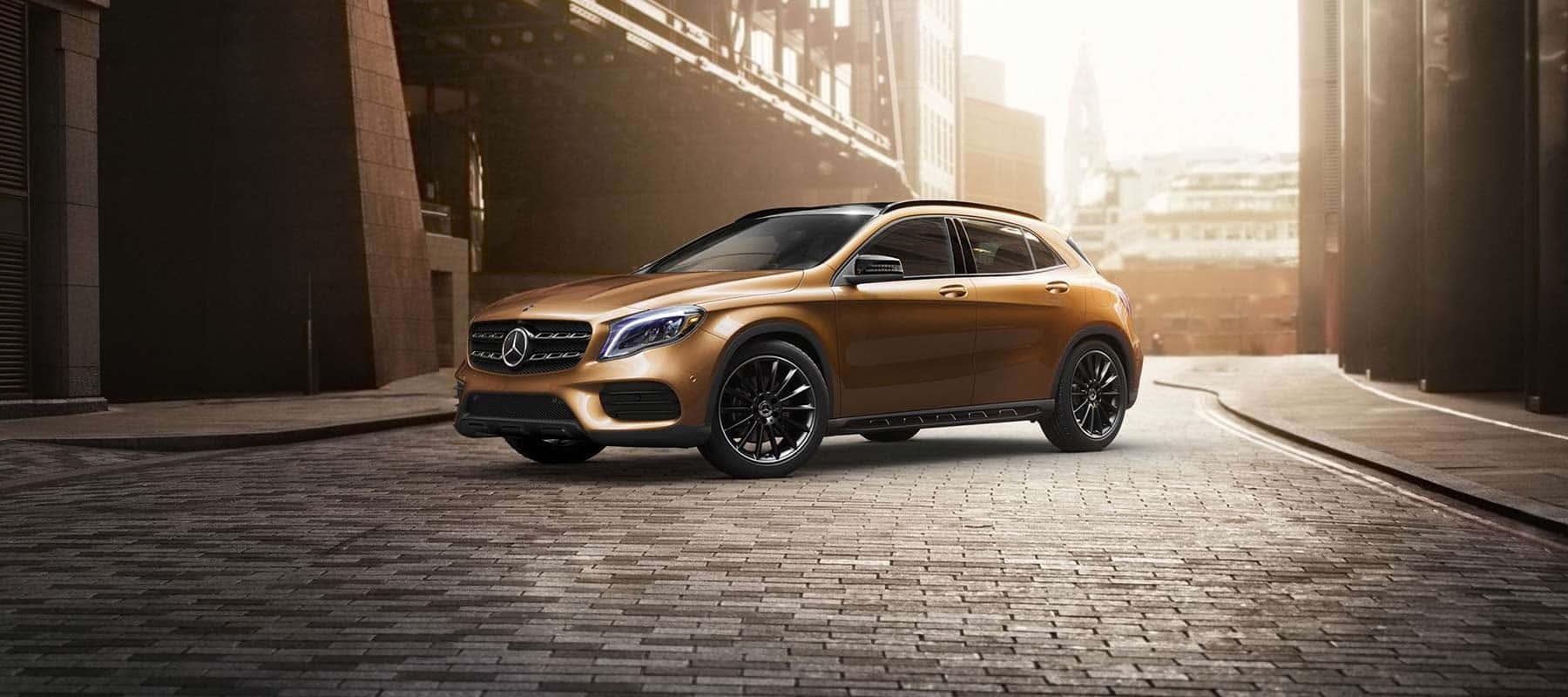 Mercedes Benz Gla-class 桌布 壁紙 — 一輛閃閃發光的金色 Mercedes-Benz GLA-Class SUV 停放在現代建築之間陽光普照的鵝卵石小巷中。