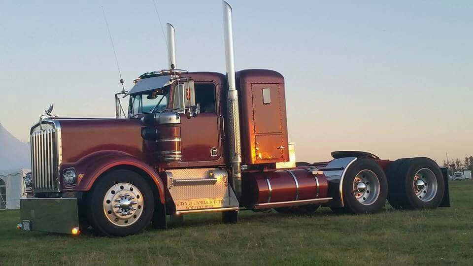 Kenworth K100 桌布 壁紙 — 一輛閃亮的栗色半掛卡車帶有鍍鉻裝飾，在日落時自豪地站在草地上，其巨大的車輪和高高的排氣管捕捉著溫暖的光線。